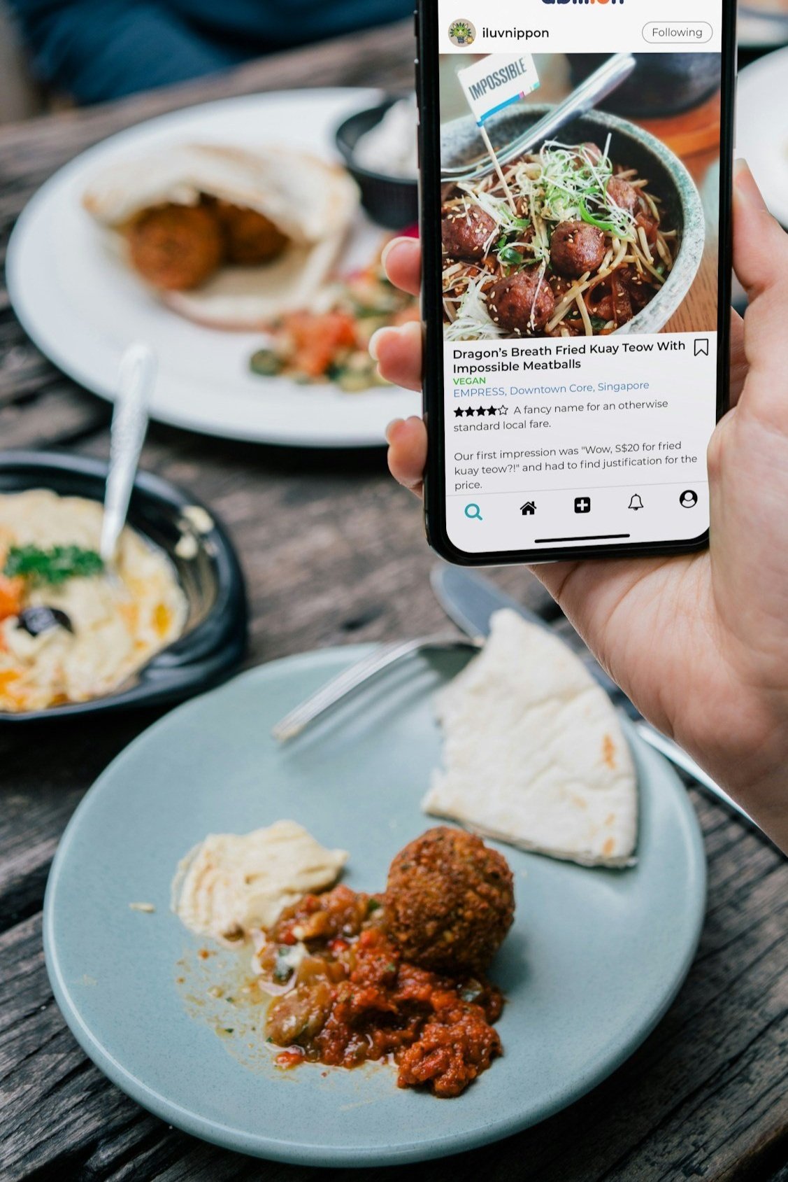 A person holding a smartphone taking a photo of a dish called fried kuay teow with meatballs, accompanied by rice and curry on a table with other dishes in the background.