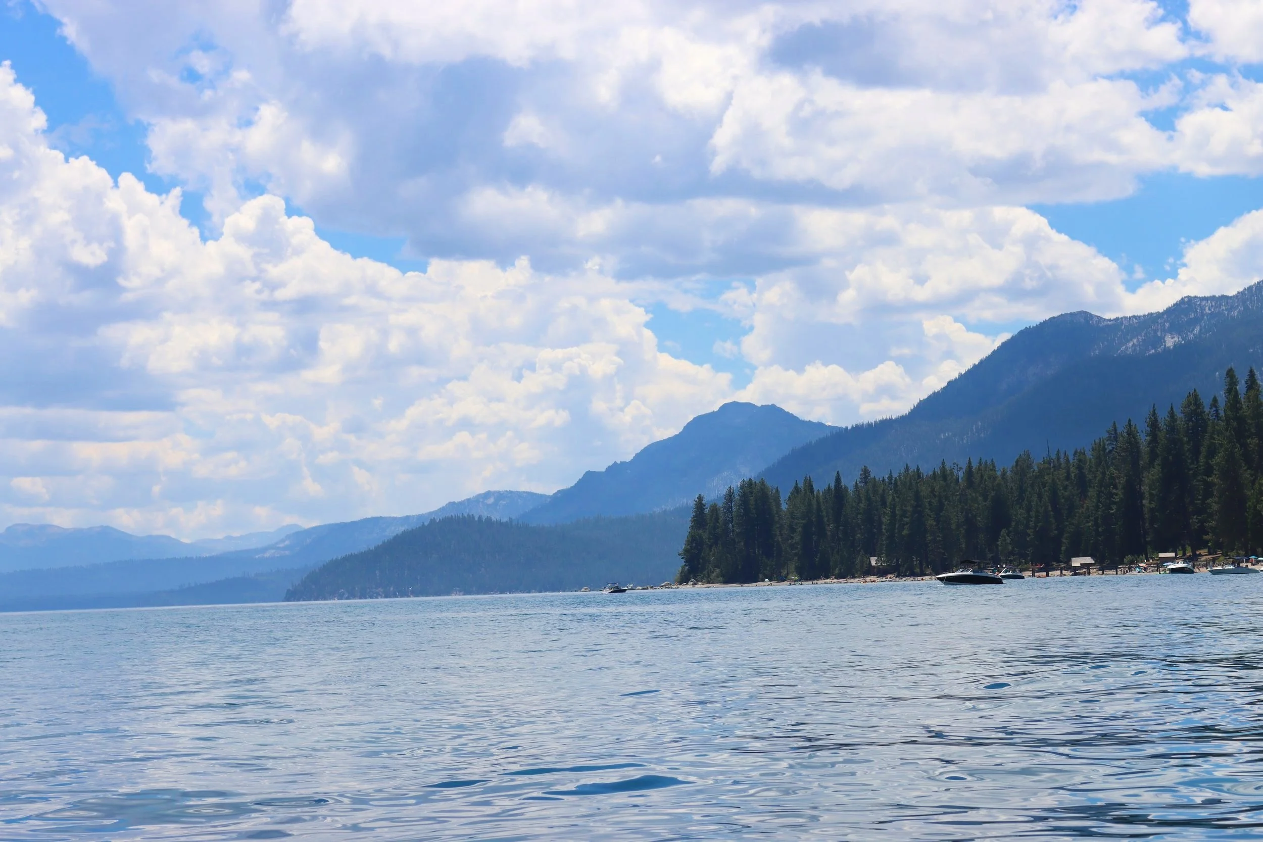 A serene lake surrounded by pine trees with mountains in the distance and a partly cloudy sky.