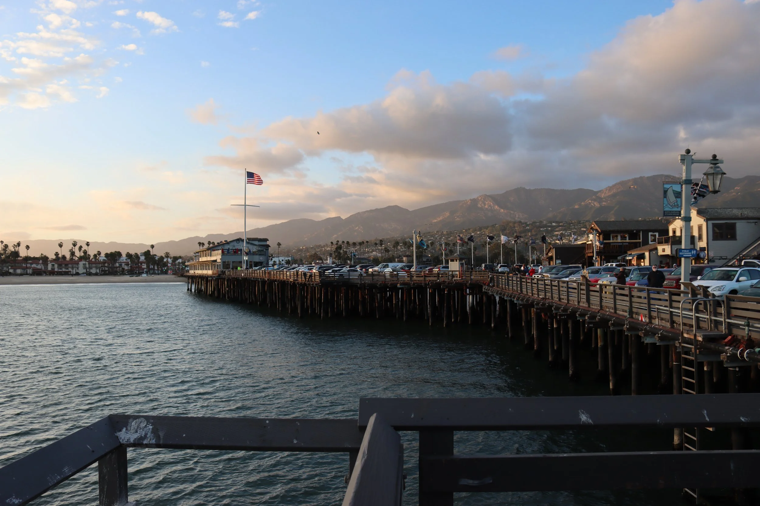 View of a pier extending over the water at sunset, with mountains and cloudy sky in the background. The pier has cars parked on it and American flags flying.