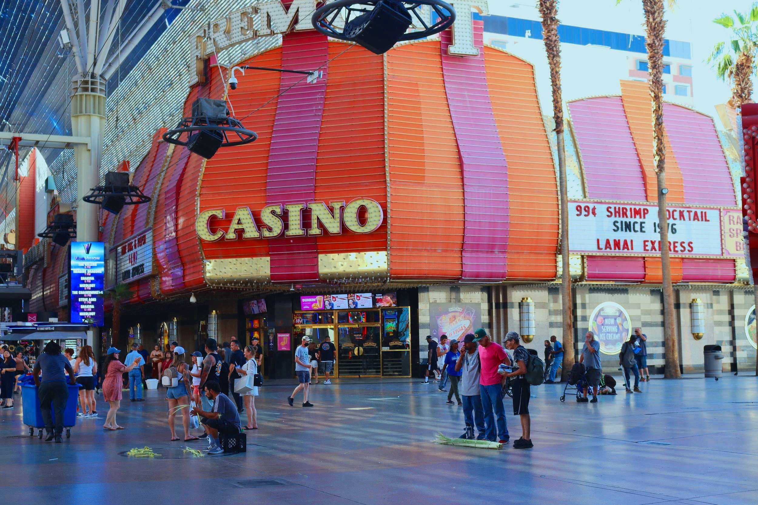 People walking and gathering outside a large, brightly lit casino building on the Las Vegas Strip, with palm trees and digital advertisements in the background.
