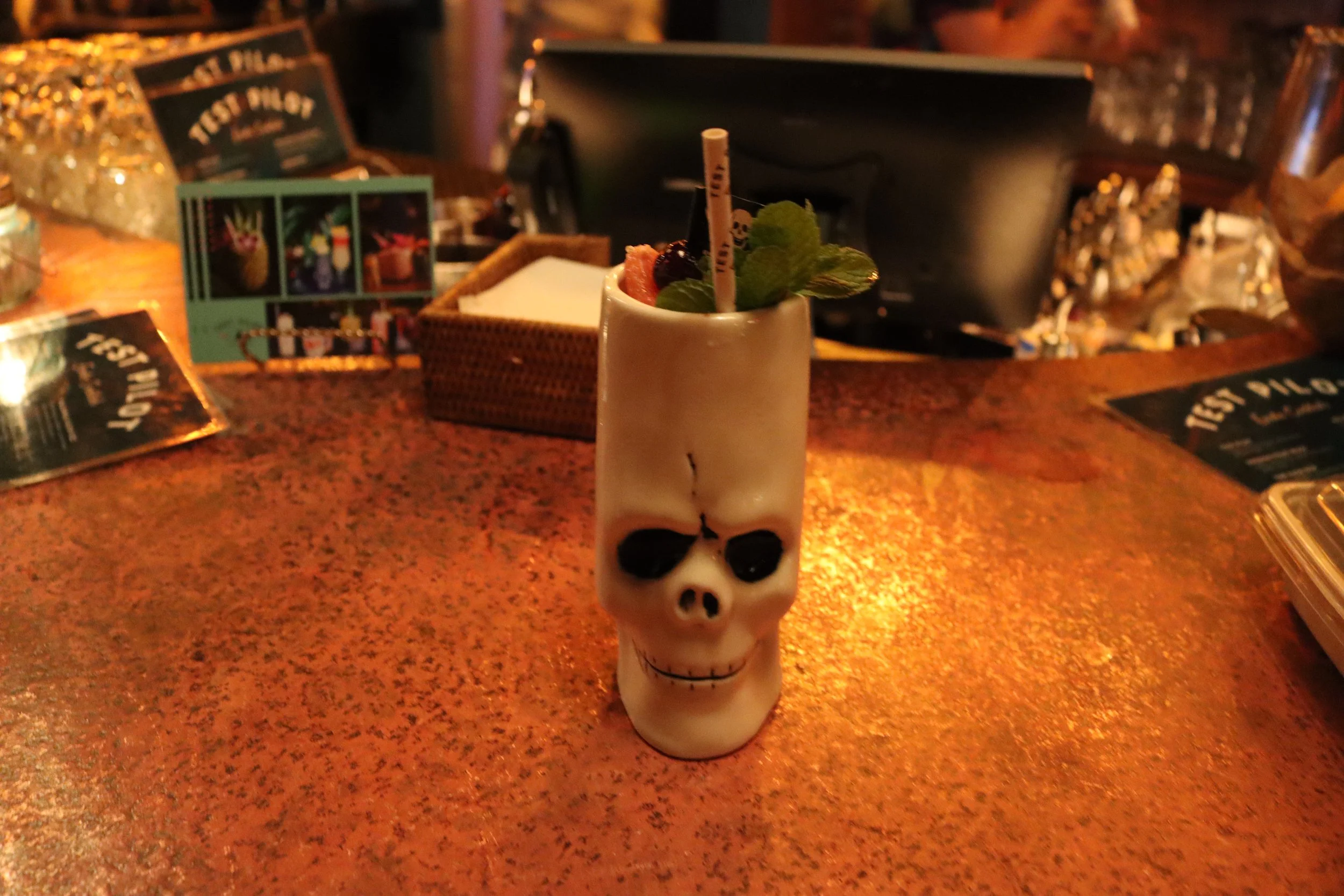 A drink in a skull-shaped mug with a black eye socket design, garnished with mint leaves, berries, and a straw, on a red speckled bar counter.