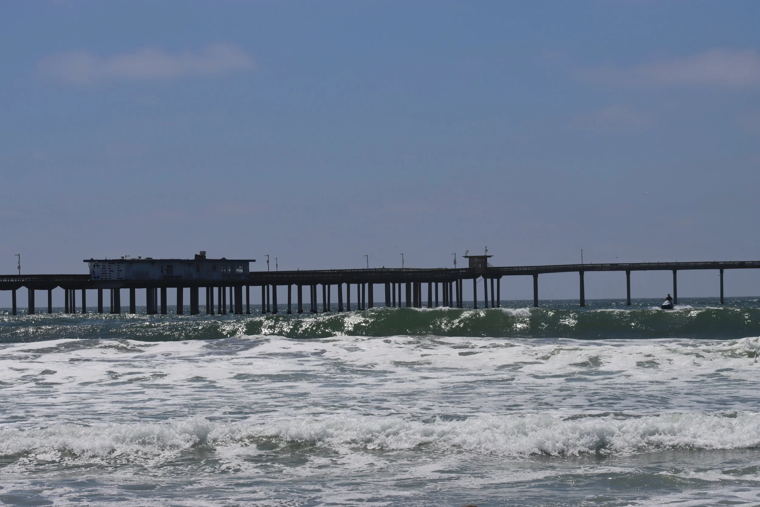 The image shows a beach scene with waves crashing onto the shore and a long pier extending into the ocean with a small building at the beginning of the pier and a person riding a jet ski off to the right.
