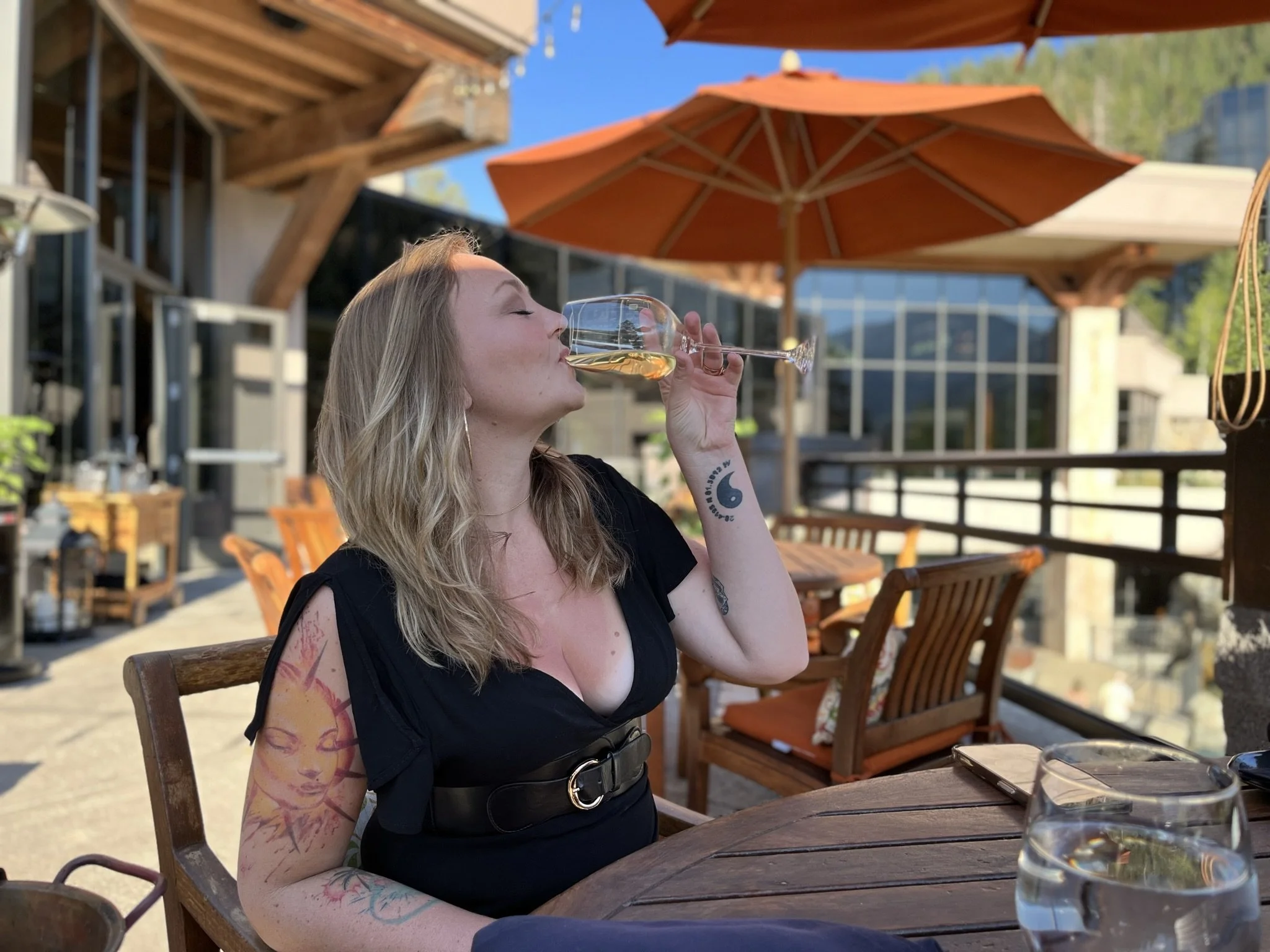A woman with tattoos and blonde hair, dressed in a black dress, is sitting at an outdoor restaurant table, drinking from a wine glass of white wine or champagne. She is seated on a wooden chair, with orange patio umbrellas and modern building glass walls in the background on a sunny day.