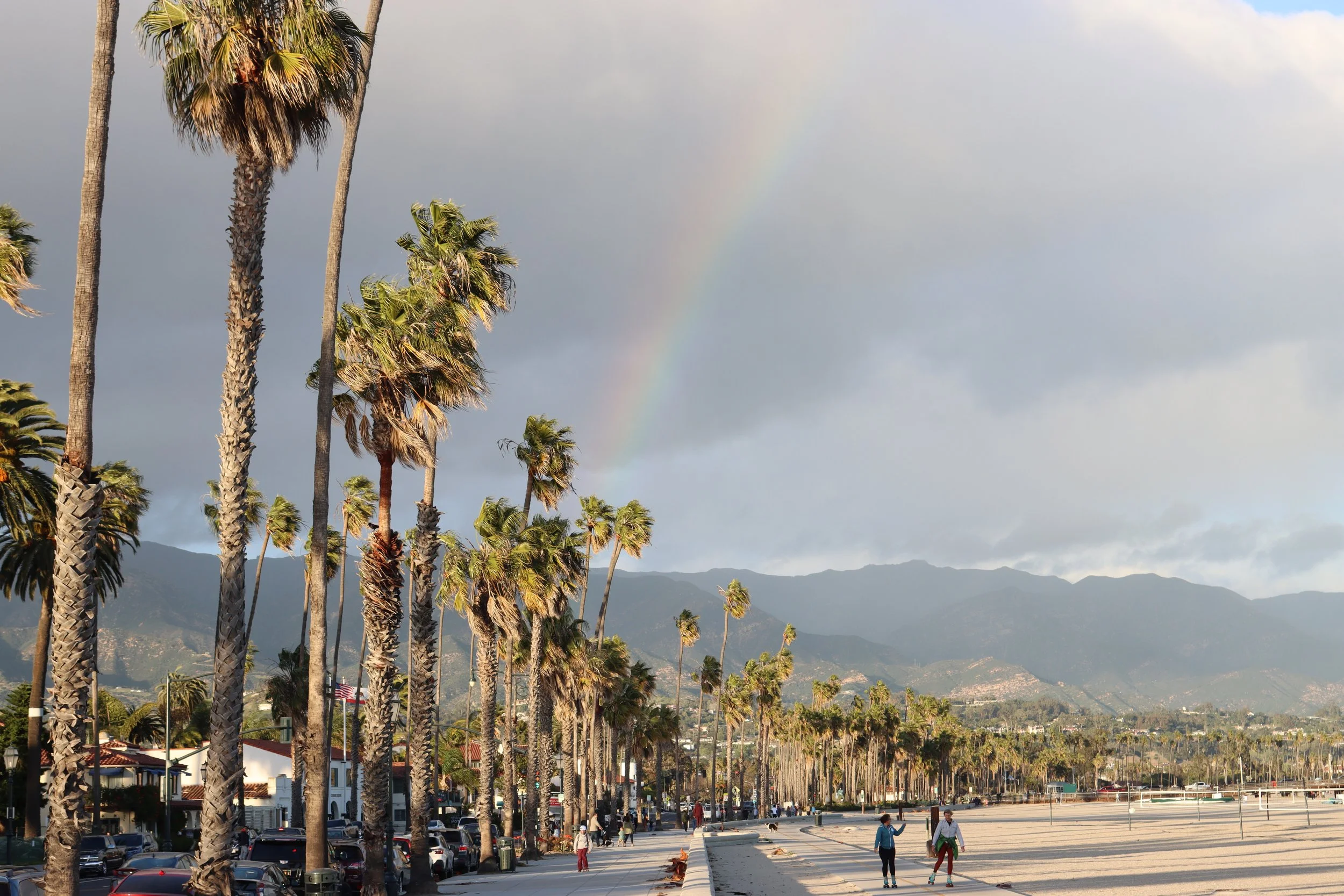 Palm trees along a sidewalk with mountains in the background, cloudy sky, faint rainbow, and people walking in Southern California.