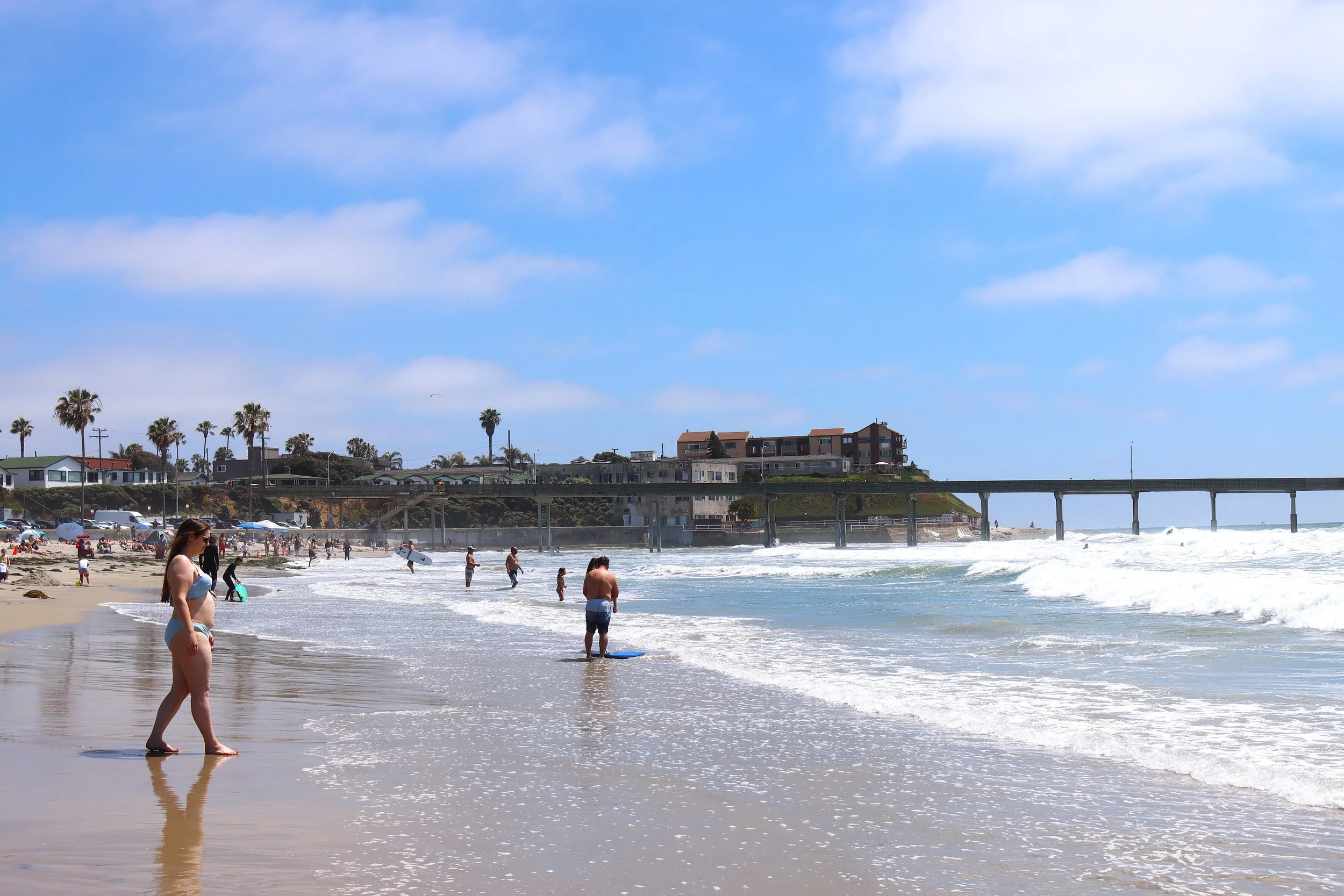 People relaxing and swimming on a sunny beach with a pier and houses on cliffs in the background.