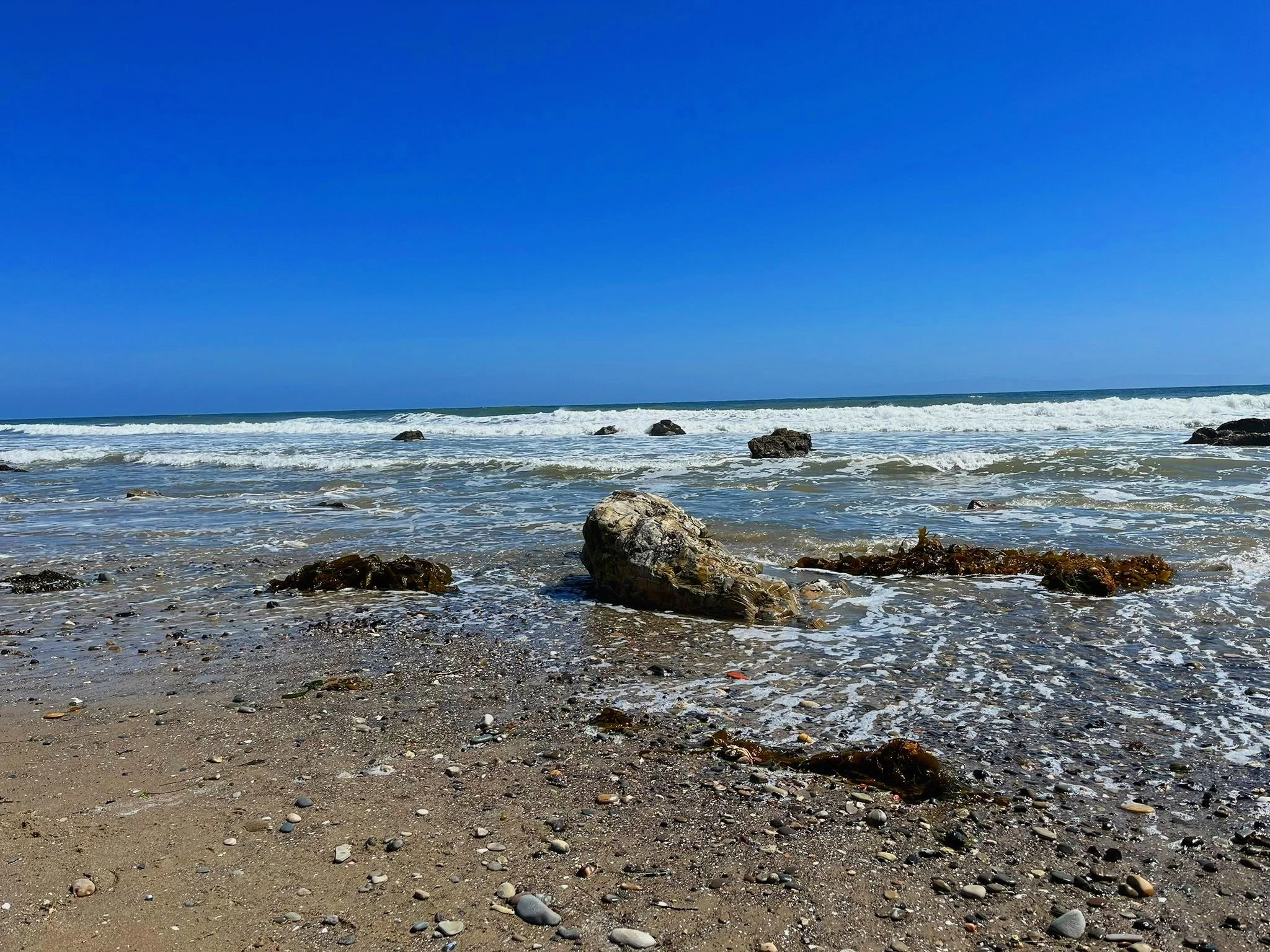 A sandy beach with scattered rocks and seaweed, ocean waves, and a clear blue sky.