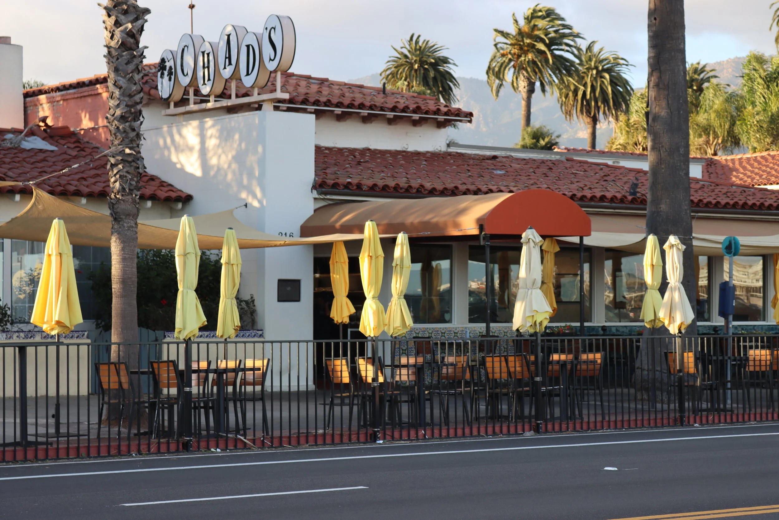 Empty outdoor patio with yellow umbrellas and wooden chairs in front of a restaurant with a sign that reads 'Chad's', palm trees, and mountainous background.