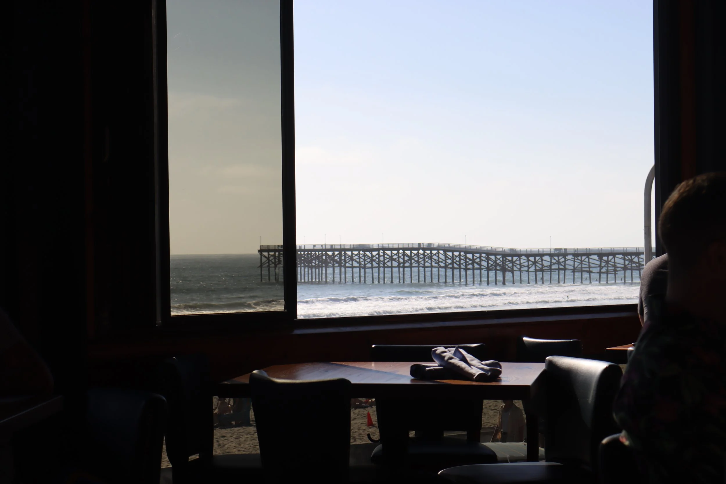 View of the ocean through a large window with a pier extending into the water, seen from inside a dimly lit restaurant or café.