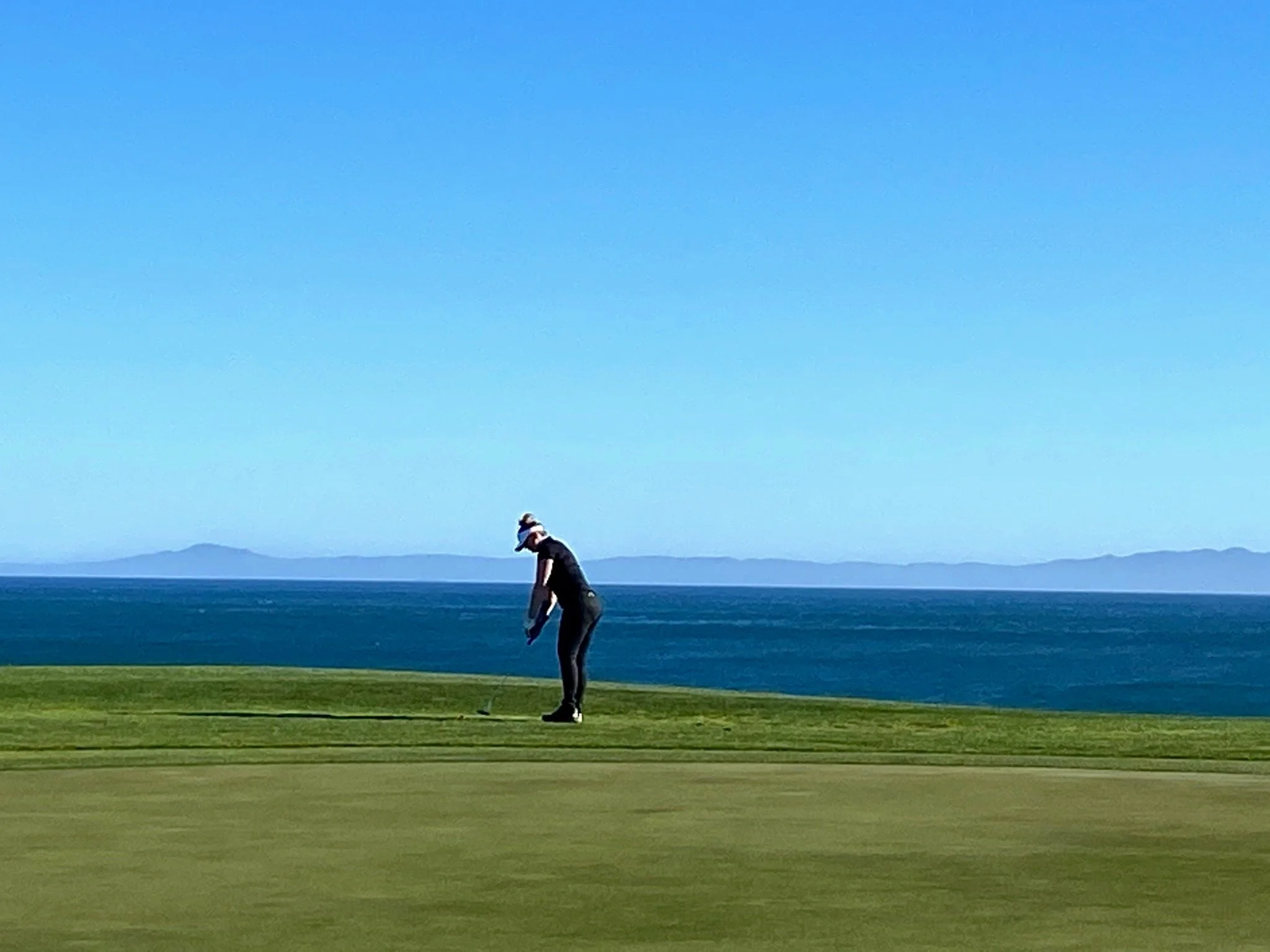 A person playing golf on a green near the ocean with a mountain range in the background.