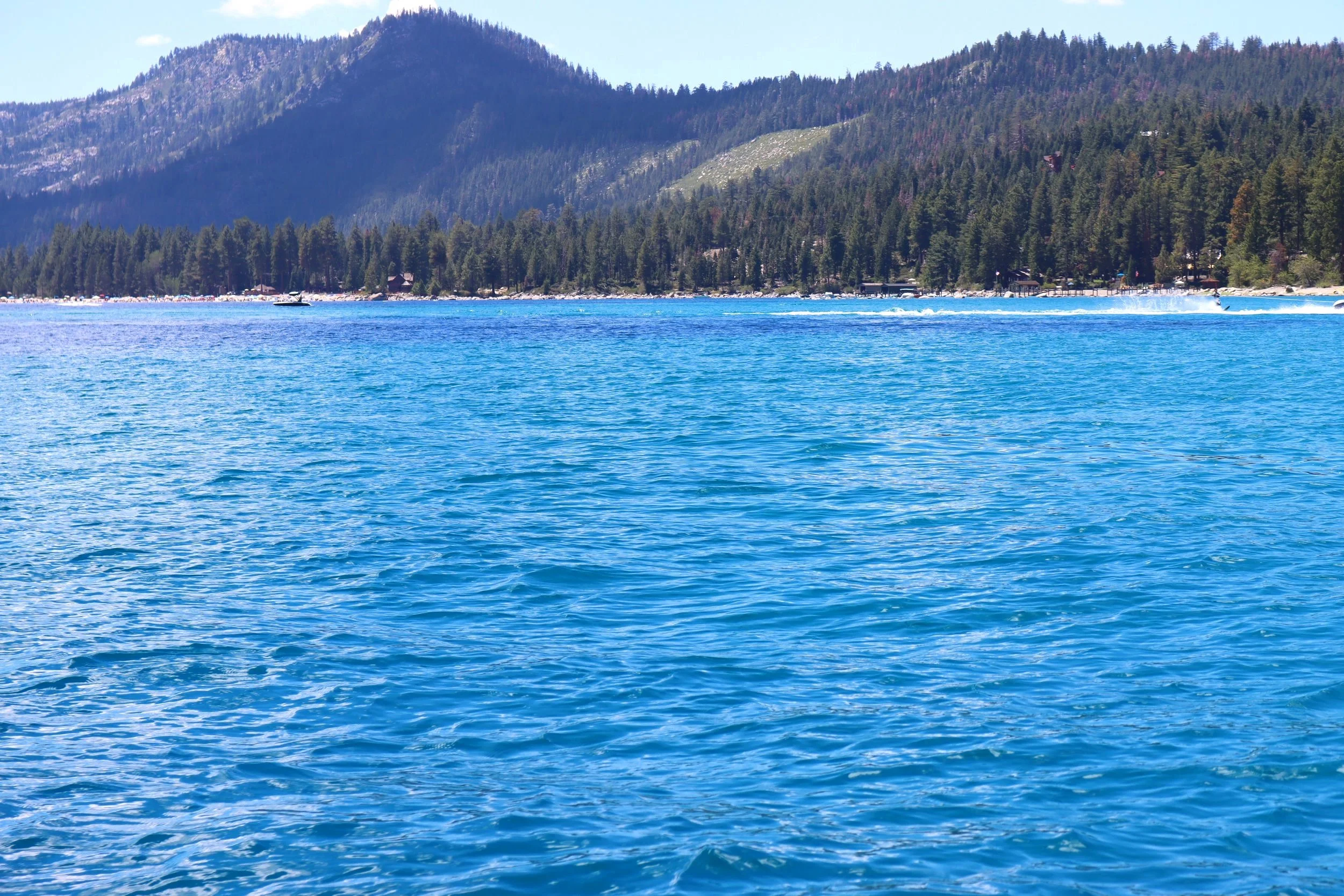 A large body of blue water with a forested mountain in the background, sunny sky.