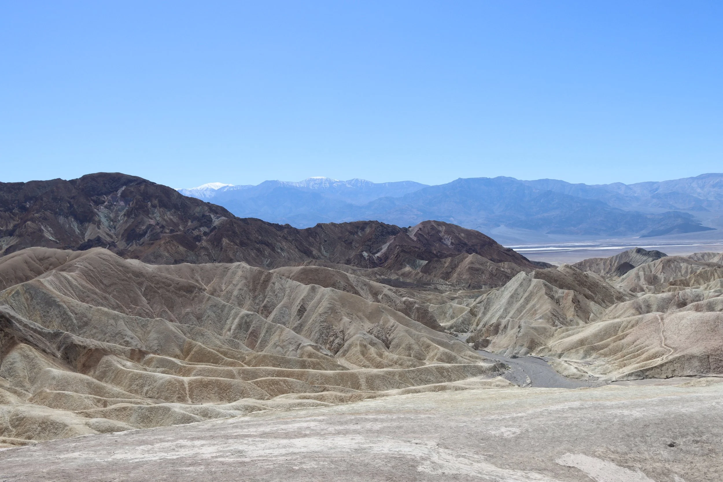 A panoramic view of desert mountains with layered, colorful ridges in the foreground, dark mountains in the middle, and snow-capped peaks under a clear blue sky in the distance.