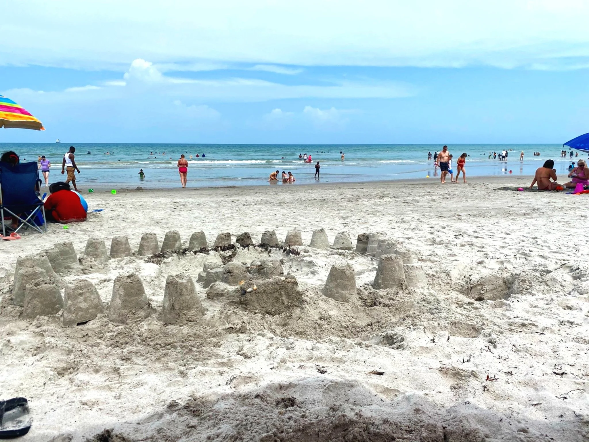 Kids sandcastle with walls and towers on sandy beach, with people swimming and relaxing by the ocean in the background on a partly cloudy day.