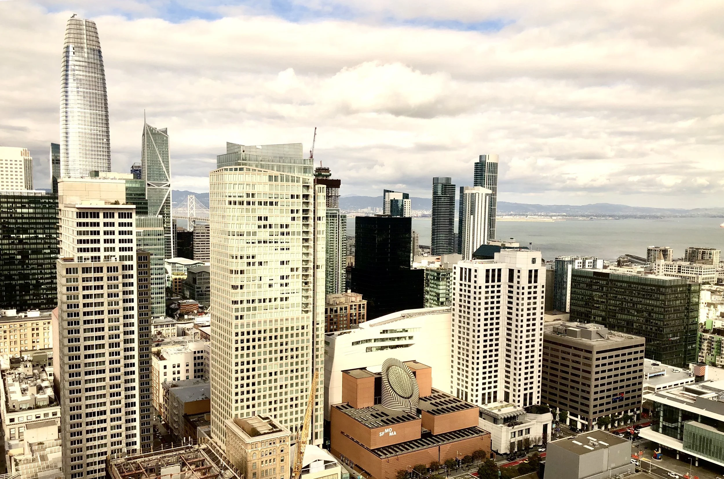 Cityscape of San Francisco with tall skyscrapers, including Salesforce Tower, and the San Francisco Bay in the background under partly cloudy skies.