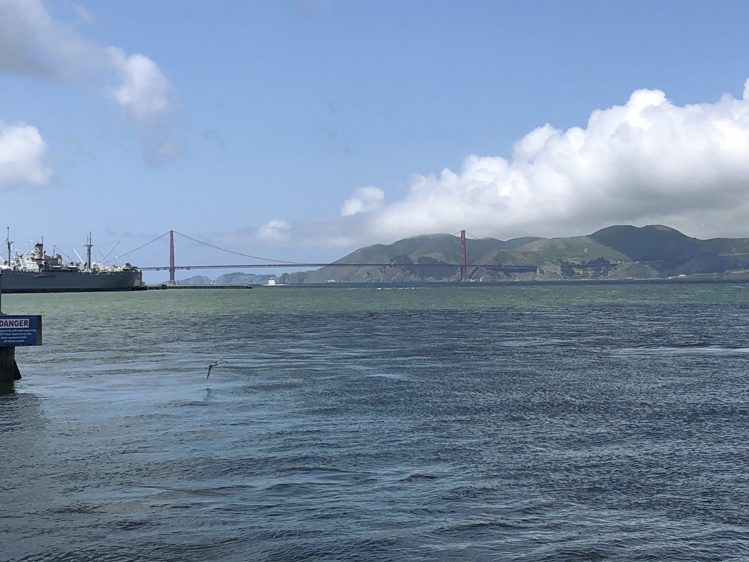 View of San Francisco Bay with the Golden Gate Bridge in the background, a ship docked on the left side, and hills under a partly cloudy sky.