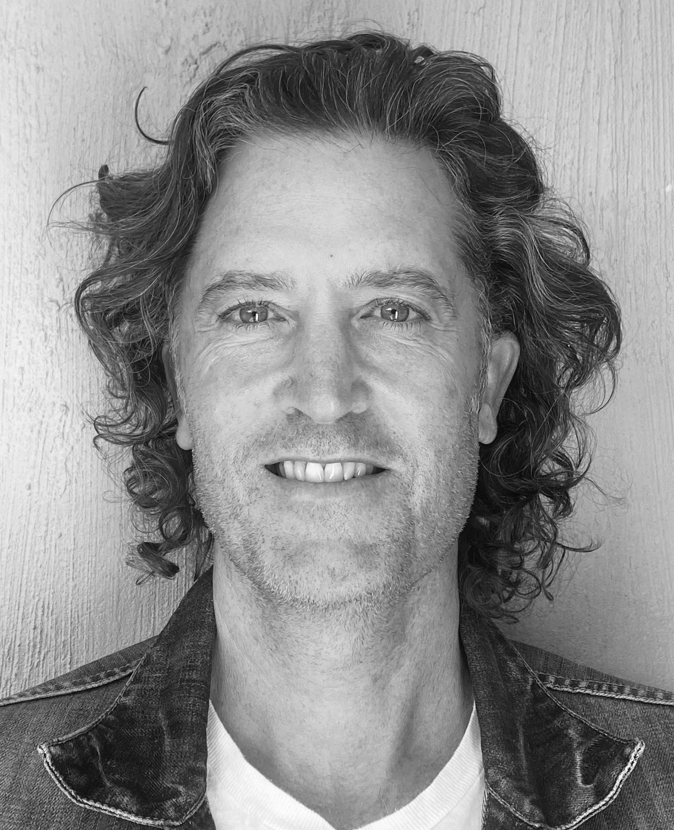 Black and white portrait of a smiling man with curly hair, wearing a denim jacket.