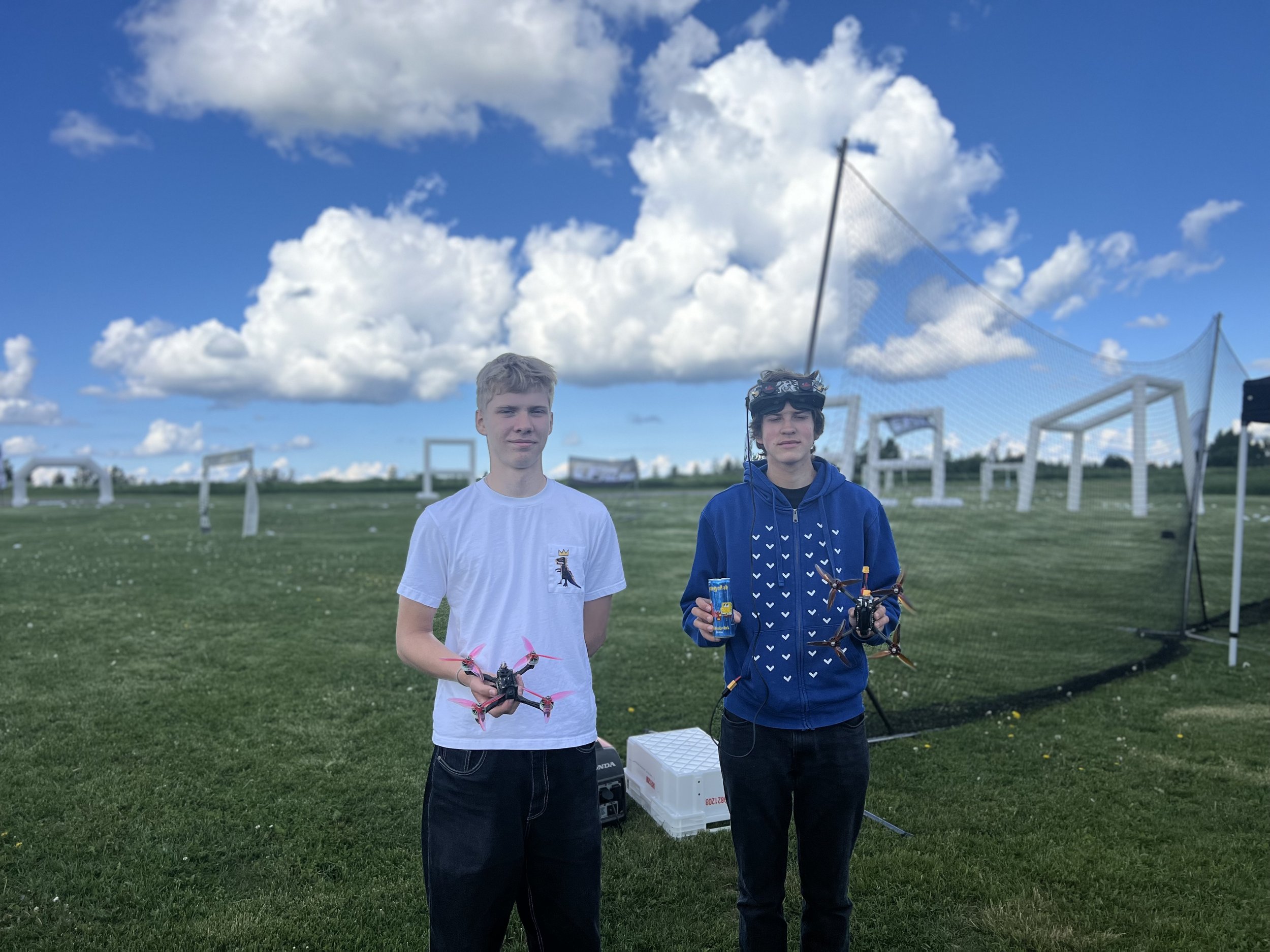 Two young men standing on a grassy field holding drones, with drone racing gates and a blue sky with clouds in the background.
