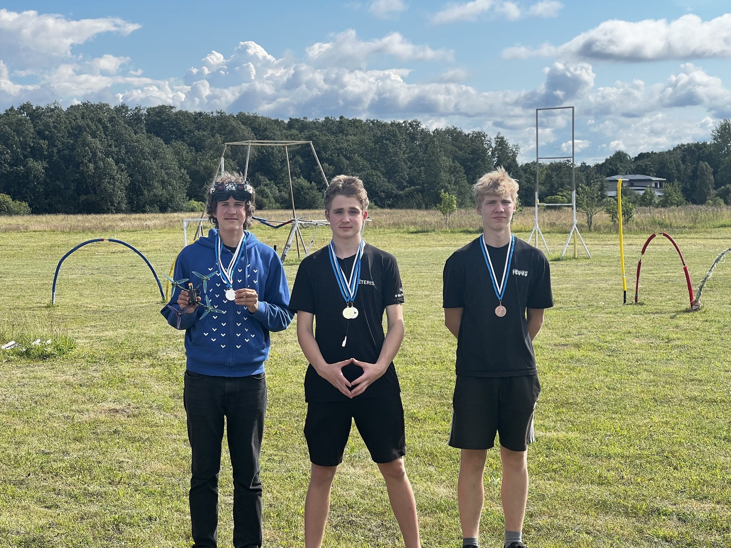 Three young men standing on a grassy field wearing medals, with one holding a drone. Background includes outdoor sports equipment and a line of trees under a partly cloudy sky.