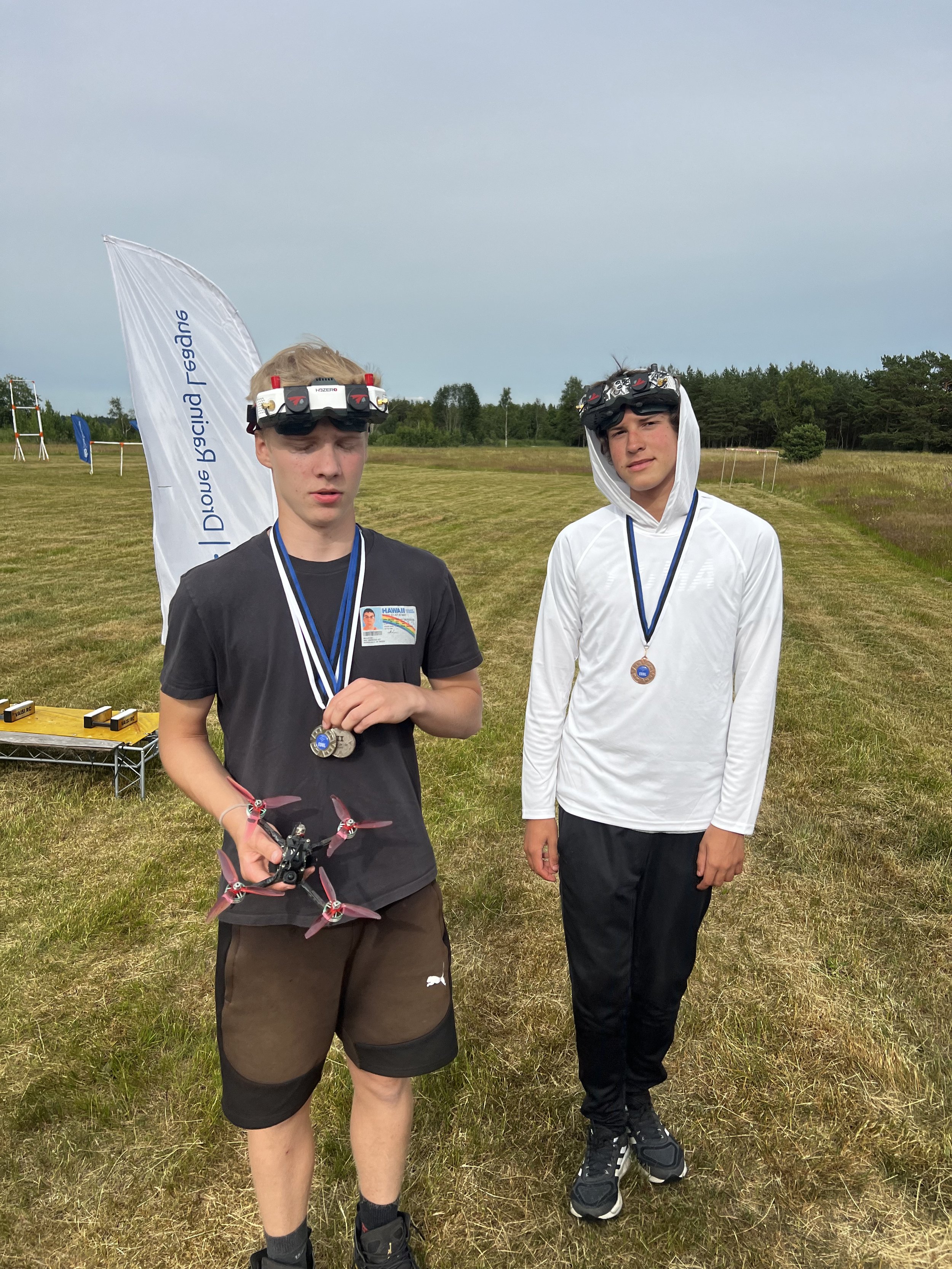 Two individuals outdoors holding medals and wearing headgear, one holding a drone. Open grassy area in the background.