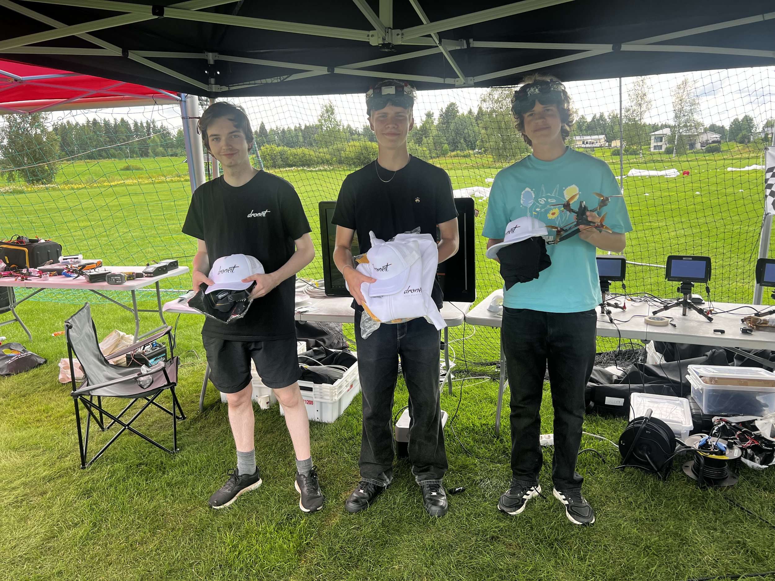 Three young men standing under a canopy holding white hats and a drone, with electronic equipment and chairs on tables behind them, outdoors on grass.