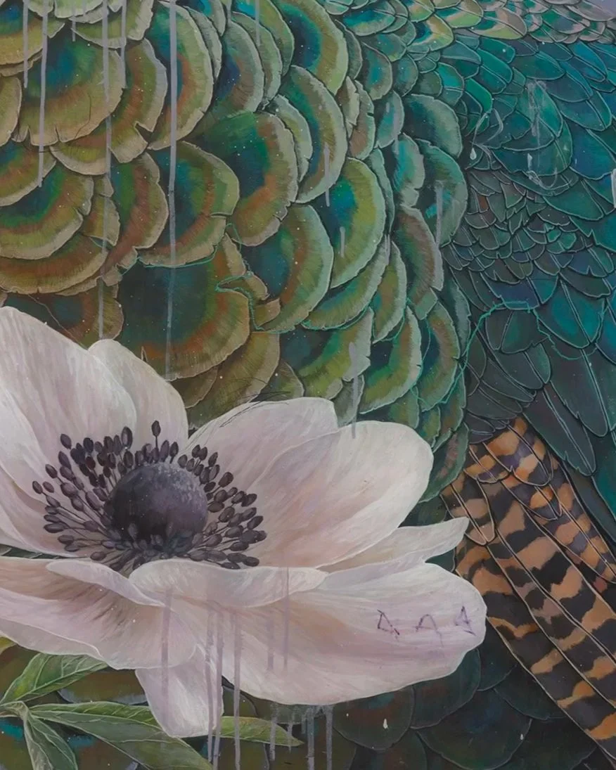 Close-up of a white anemone flower with black center, set against a background of layered green, blue, and orange patterned feathers and leaves.