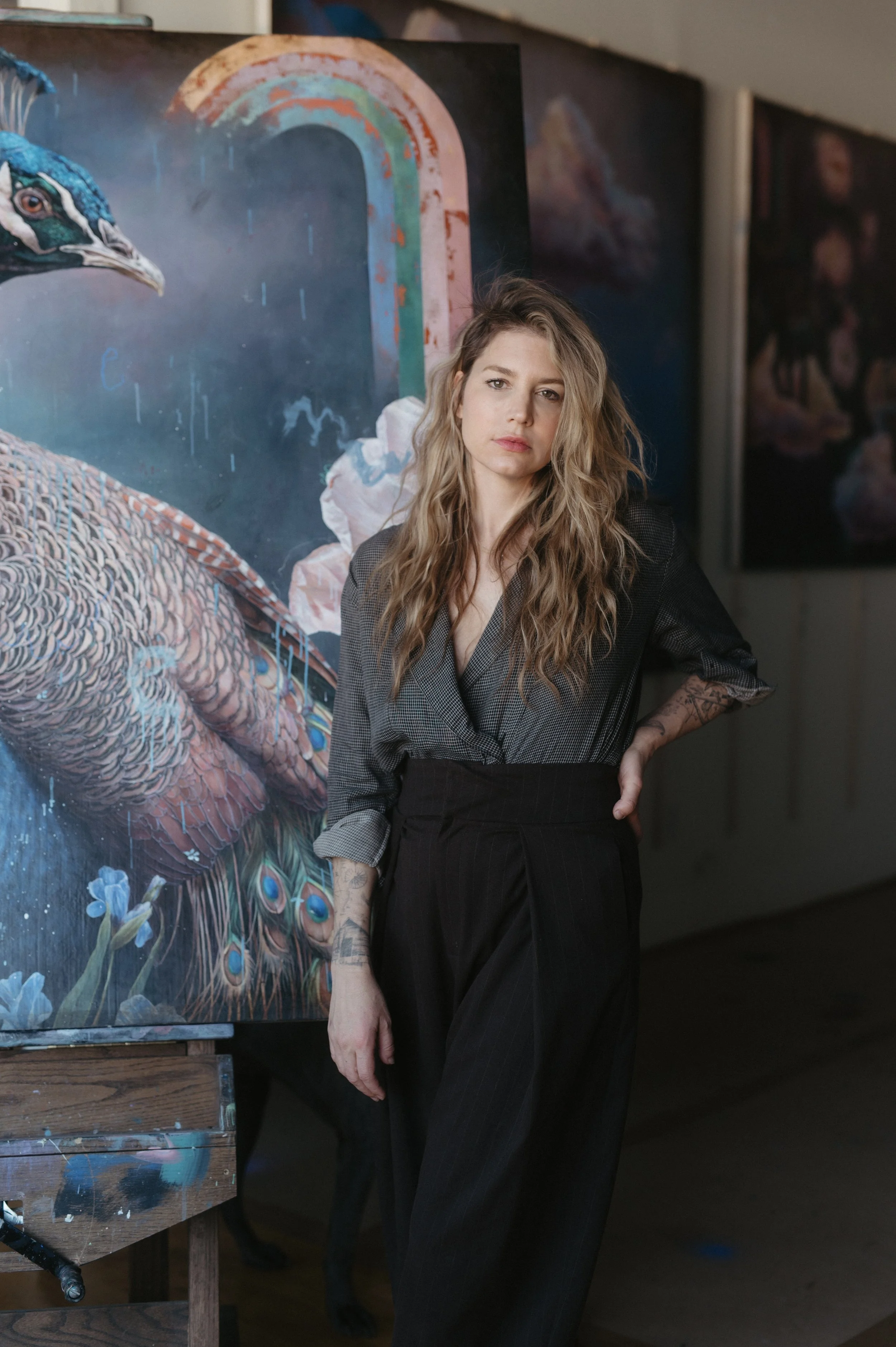 A woman with wavy blonde hair stands in front of a colorful peacock painting in an art gallery, wearing a grey checked blazer and black high-waisted pants.