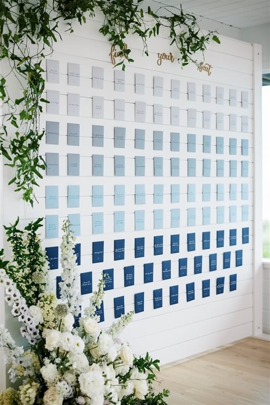 Elegant blue seating card display wall adorned with fresh white floral arrangements for a wedding