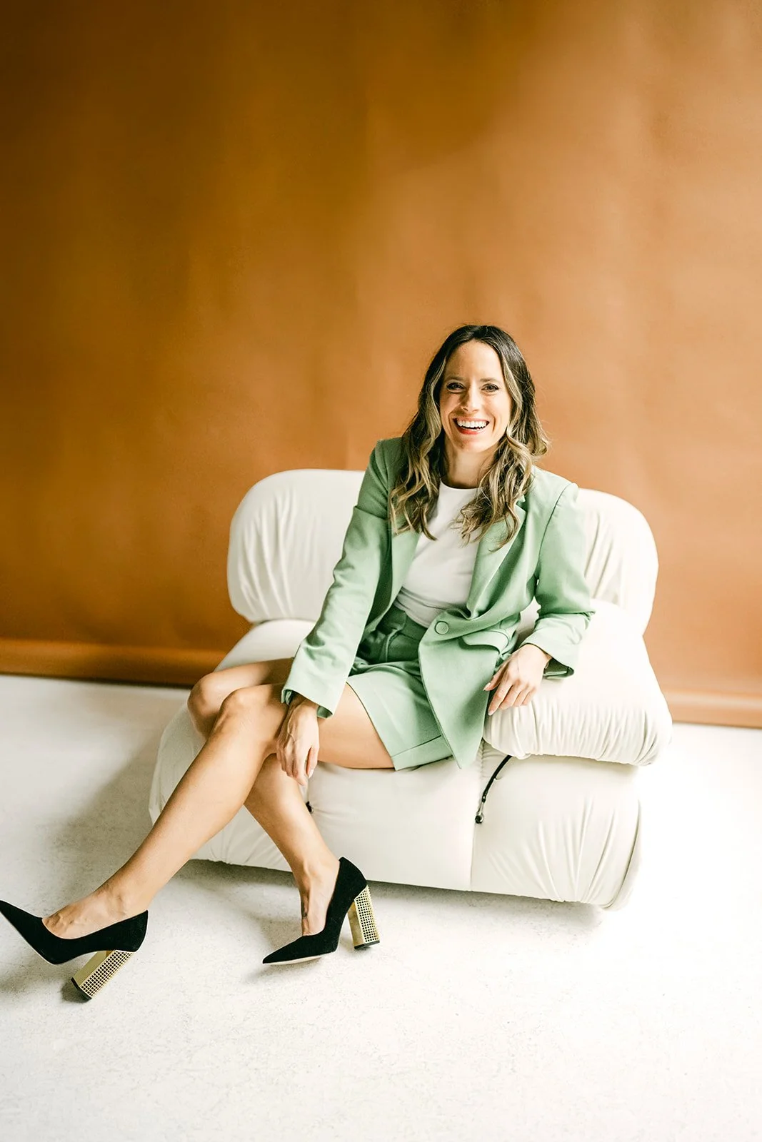 Ashley Peraino, a renowned NYC wedding planner, is pictured with a warm smile, sitting in a photo studio, her expertise and joyful spirit captured in this personal portrait