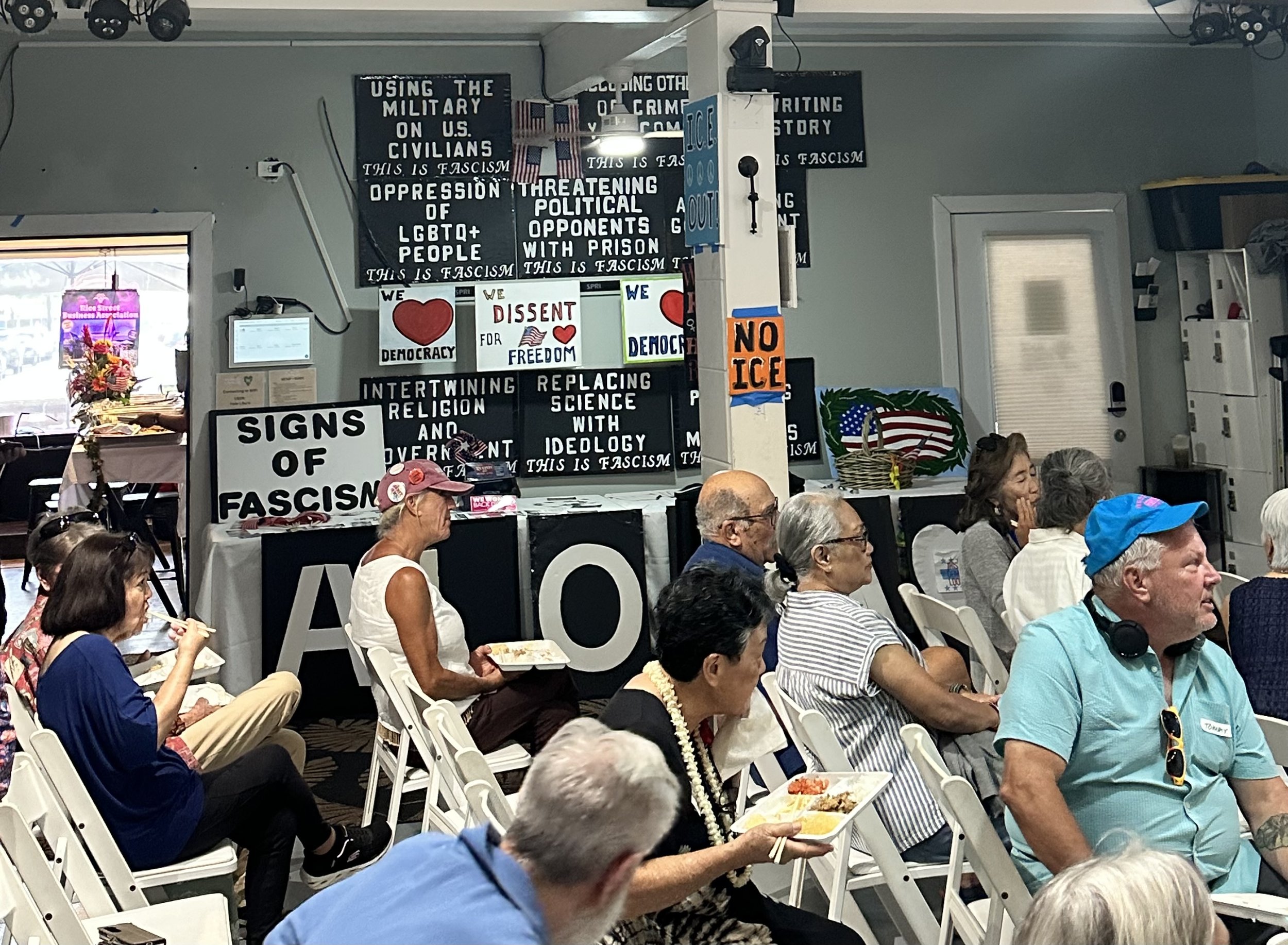 People seated in white chairs at an indoor event, with political and social signs and banners on the wall, including messages protesting fascism, advocating democracy, and supporting LGBTQ+ rights.
