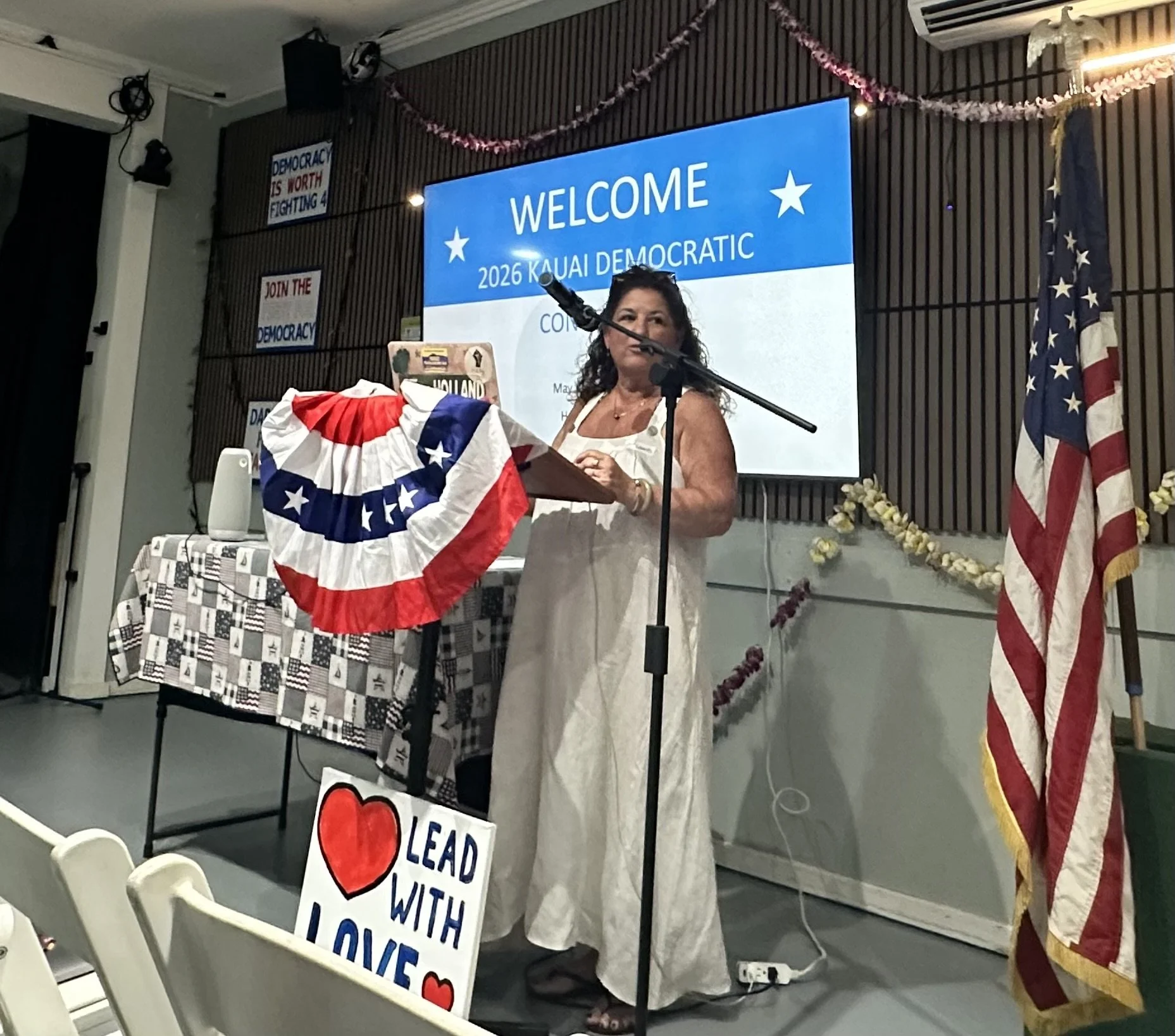 A woman standing at a podium with a microphone, decorated with a red, white, and blue bunting, at a Democratic event. Behind her is a large screen that reads 'Welcome 2026 Kauai Democratic,' and an American flag is visible to the right. Additional si