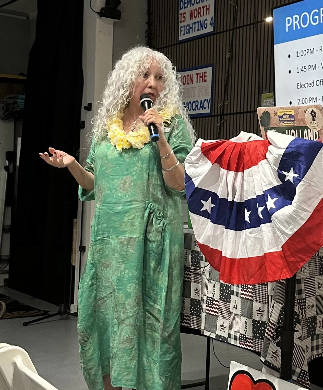 A woman with curly white hair wearing a green tie-dye dress and a yellow lei, holding a microphone and speaking at an event decorated with patriotic bunting and political signs.