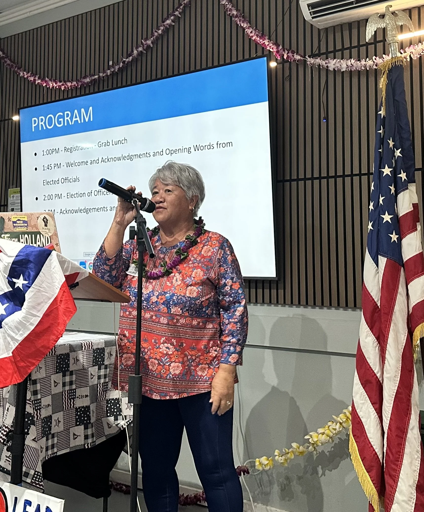 An elderly woman with gray hair, wearing a red and blue floral blouse, speaking into a microphone at a program event. Behind her is a large screen displaying the program schedule, a US flag, and a decorative lei around her neck.