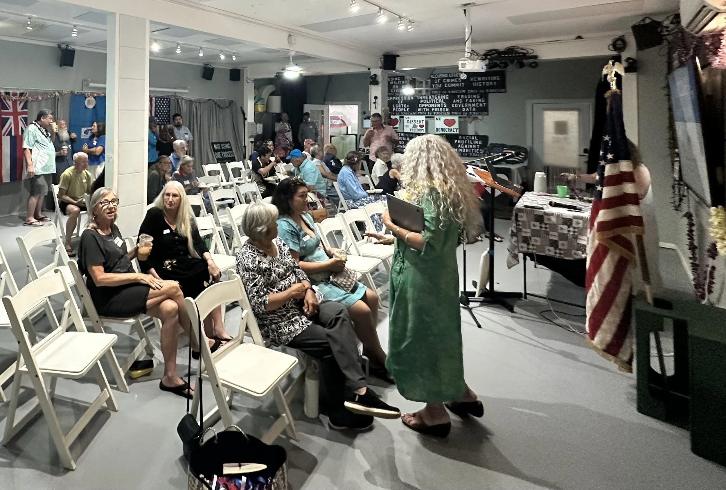 A group of diverse people seated and standing in a community room, some engaging in conversation while others are listening. There are American flags and black boards with messages on the walls