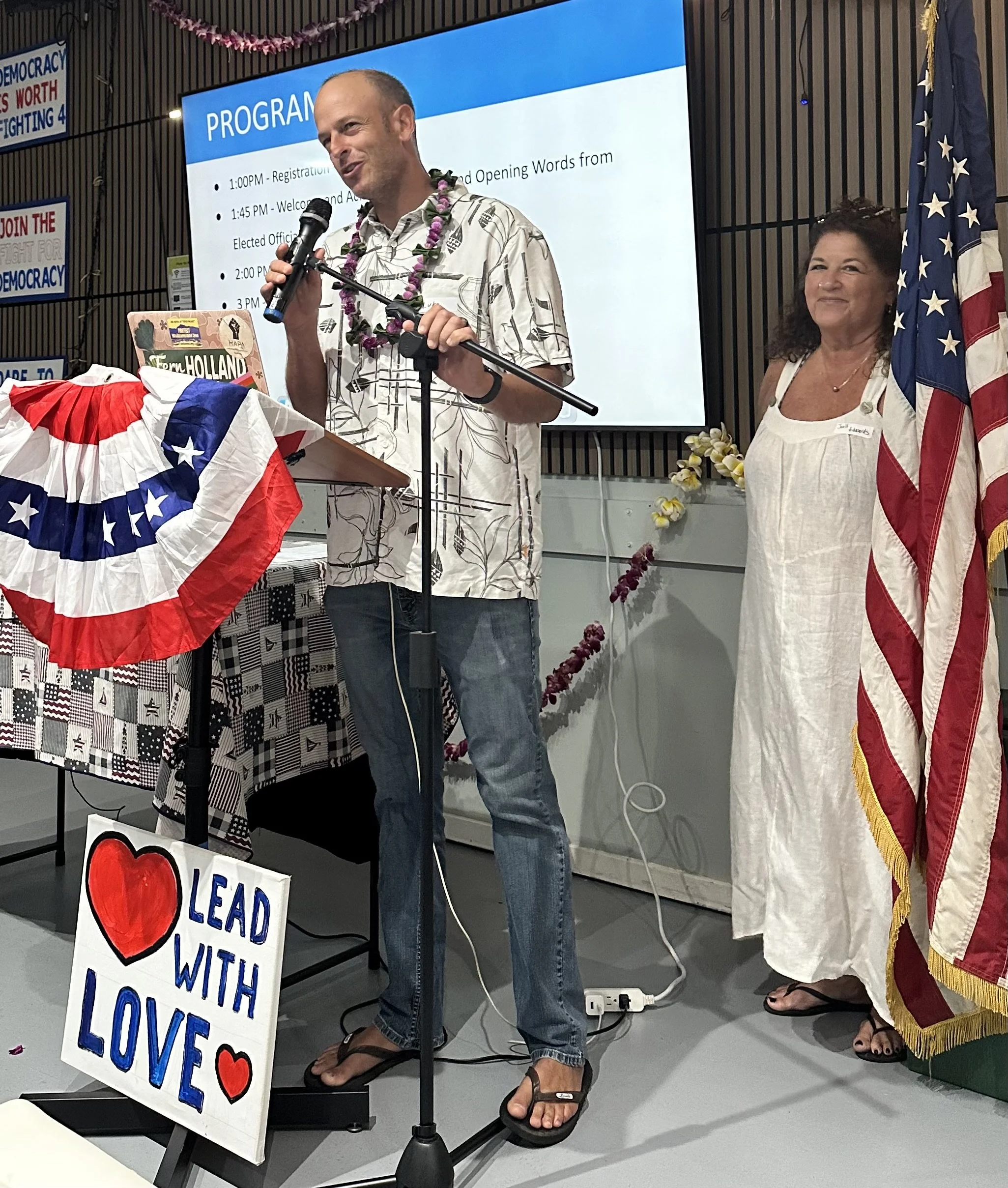 A man wearing a floral shirt and a lei, speaking into a microphone at a podium decorated with patriotic red, white, and blue bunting, at an indoor event with a large screen displaying a program schedule behind him. A woman in a white dress standing t