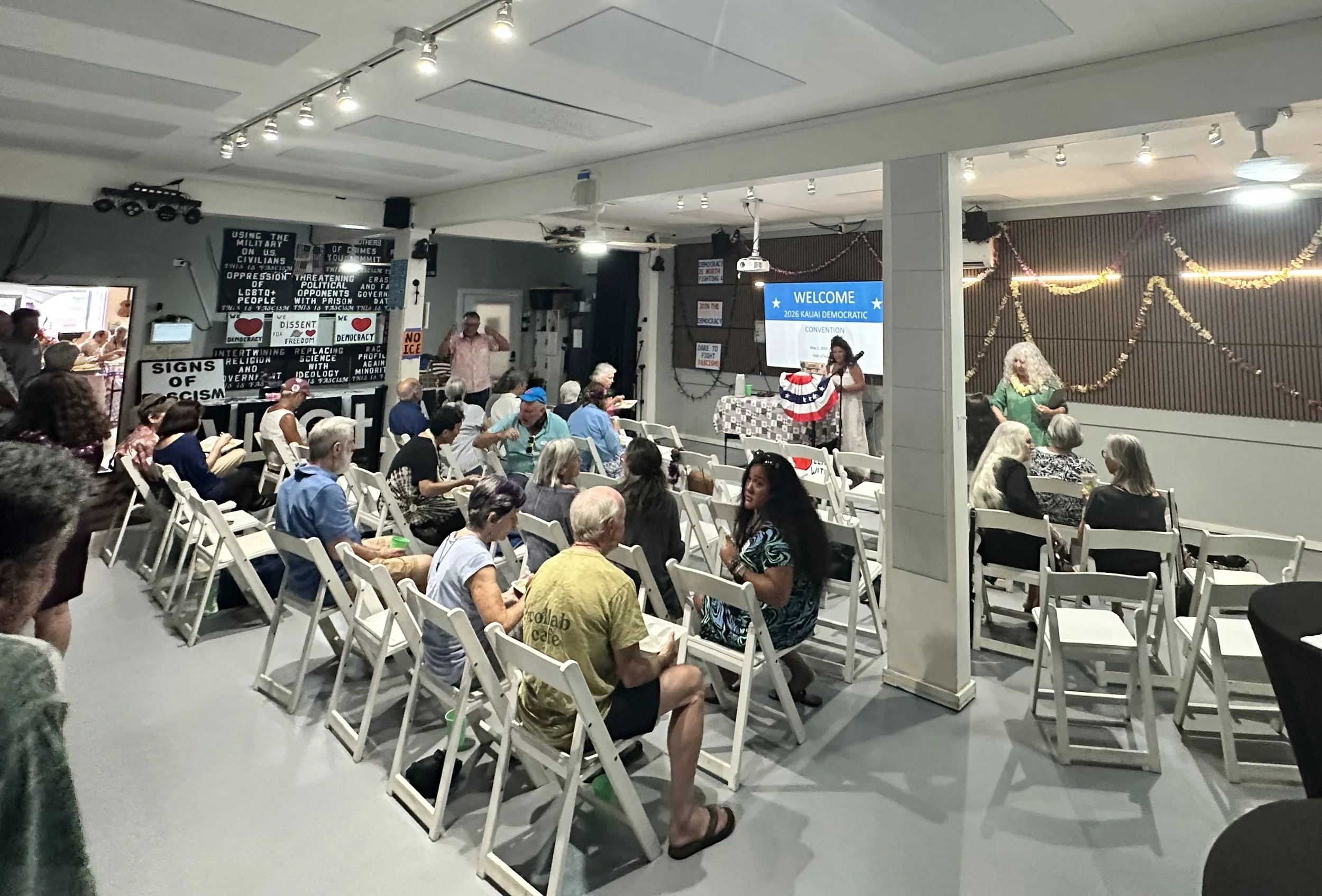 A group of people attending a community event or meeting, seated on white folding chairs in a spacious room. There is a woman standing at a podium with a large screen that reads 'WELCOME 2026 KAUI DEMOCRATIC CONVENTION.' The background behind the wom
