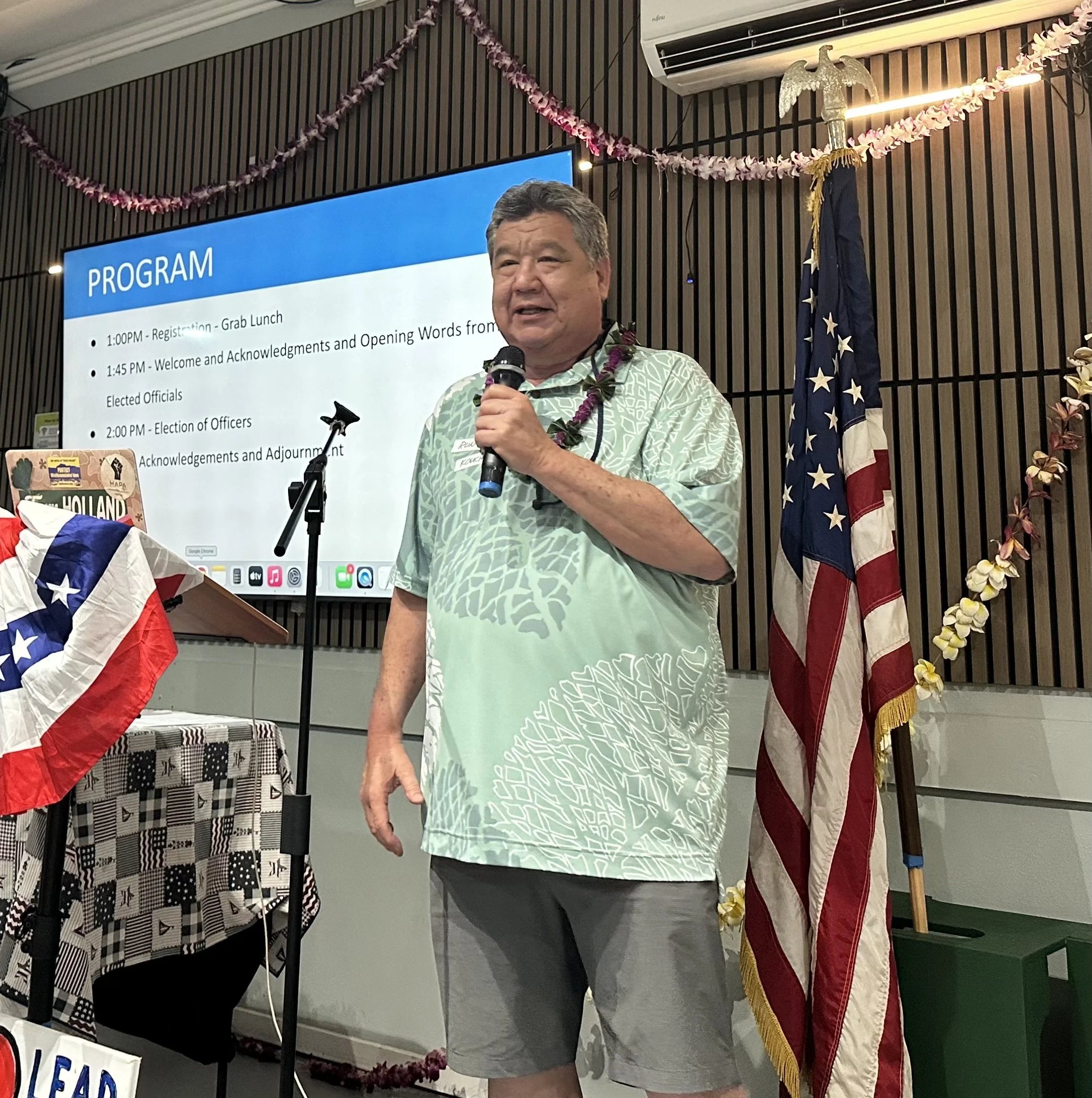 A man speaking into a microphone during a presentation at a conference, with a program slide on the screen behind him and American flags decorating the stage.