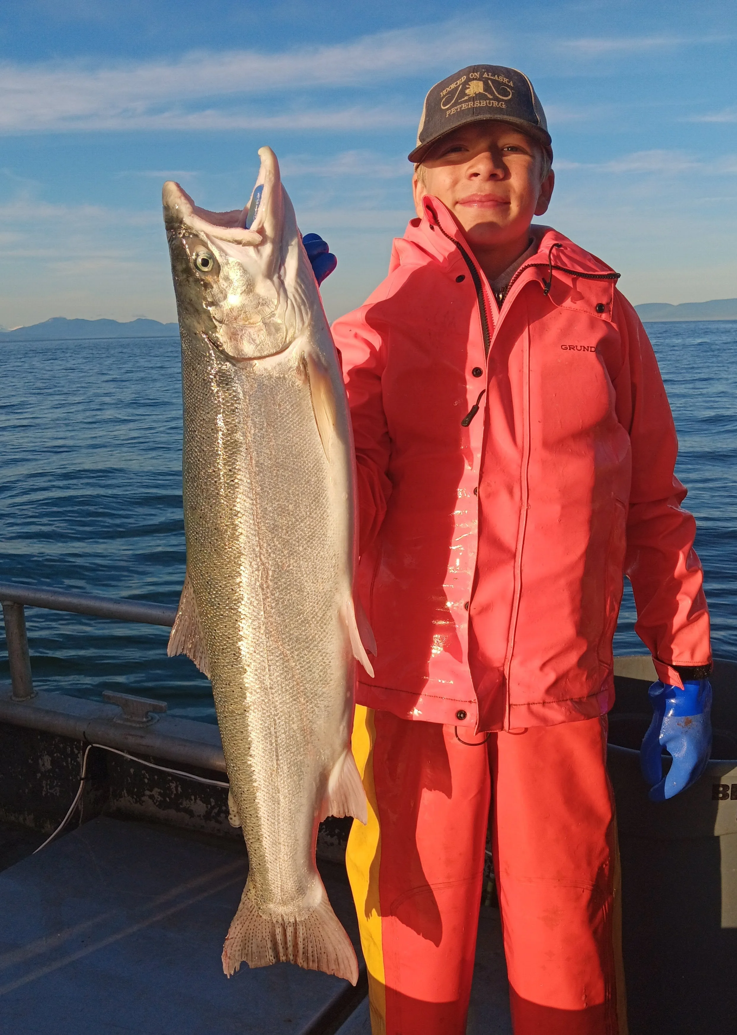 A young boy in red waterproof gear holding a large fish on a boat with the ocean and mountains in the background.