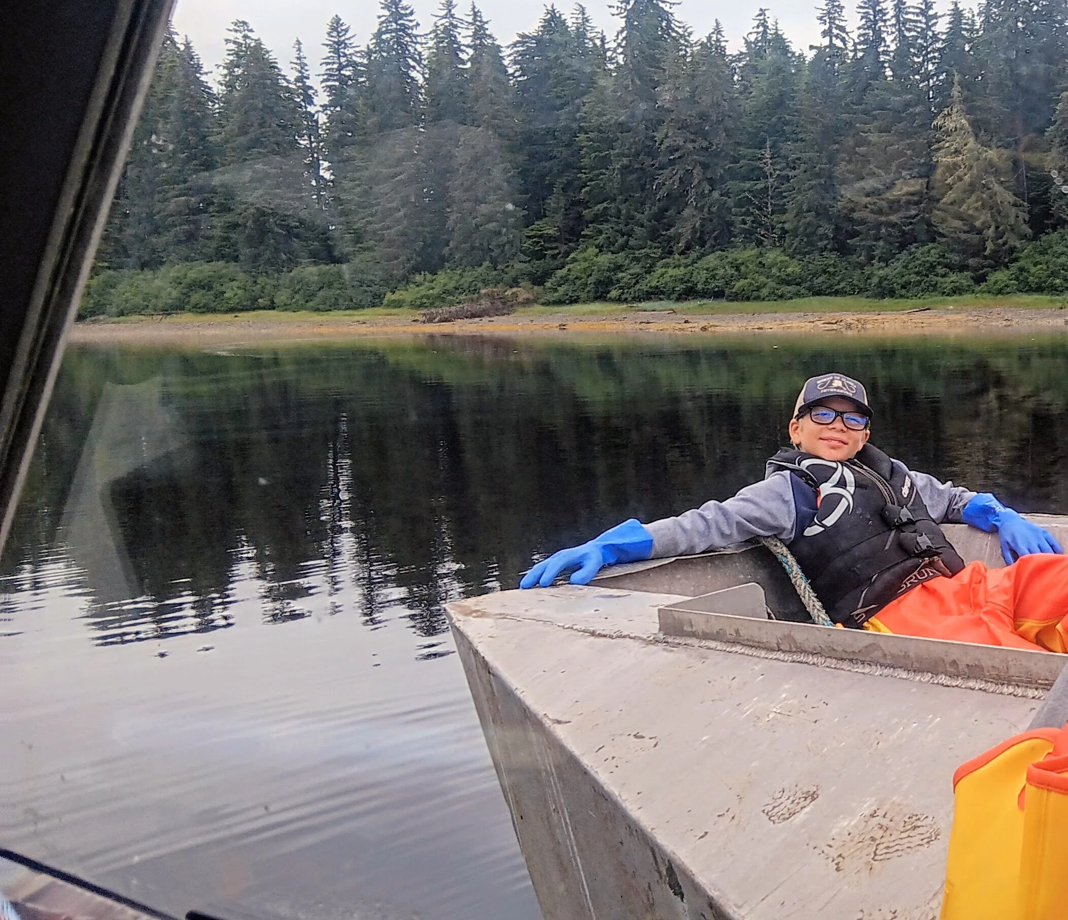 A boy wearing glasses, blue gloves, a life jacket, and orange pants is sitting in a boat on a calm body of water, smiling and relaxing with one arm draped over the side of the boat. In the background, there are trees along the shoreline.