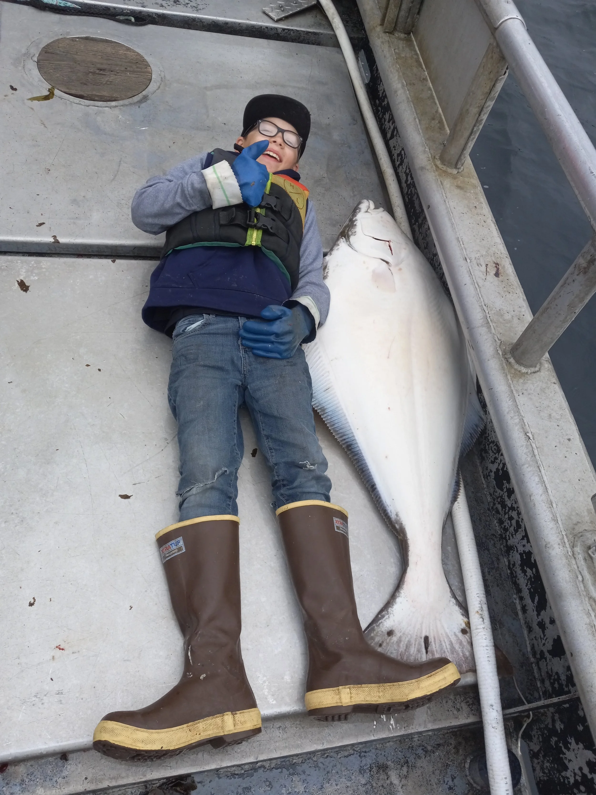 A boy lying on a boat deck next to a large, caught fish, wearing safety goggles, a life jacket, blue gloves, and brown rubber boots, smiling and giving a thumbs-up.