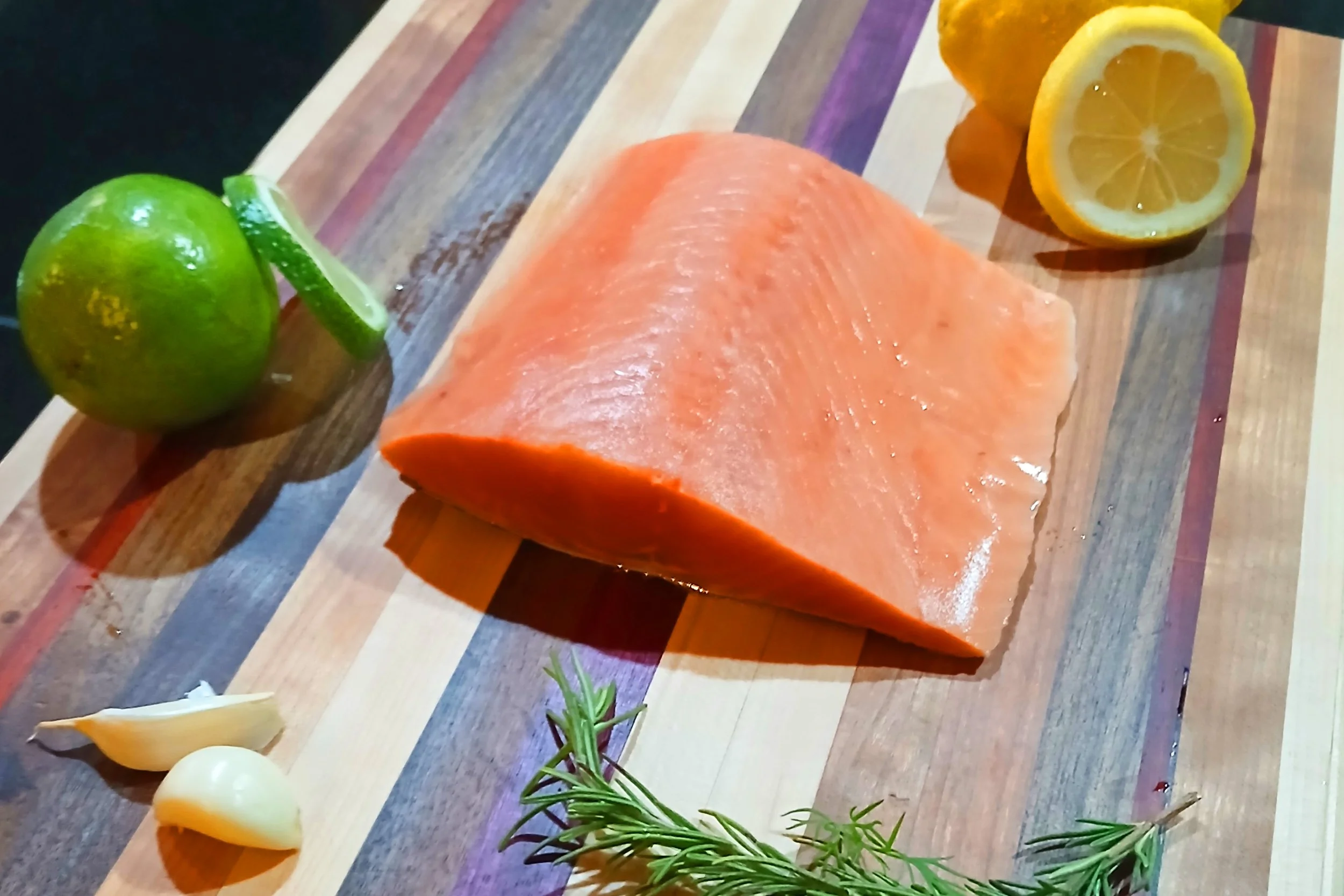 Raw salmon fillet on a colorful wooden cutting board with lemon halves, garlic cloves, a sprig of rosemary, a lime with a slice, and a lemon wedge.