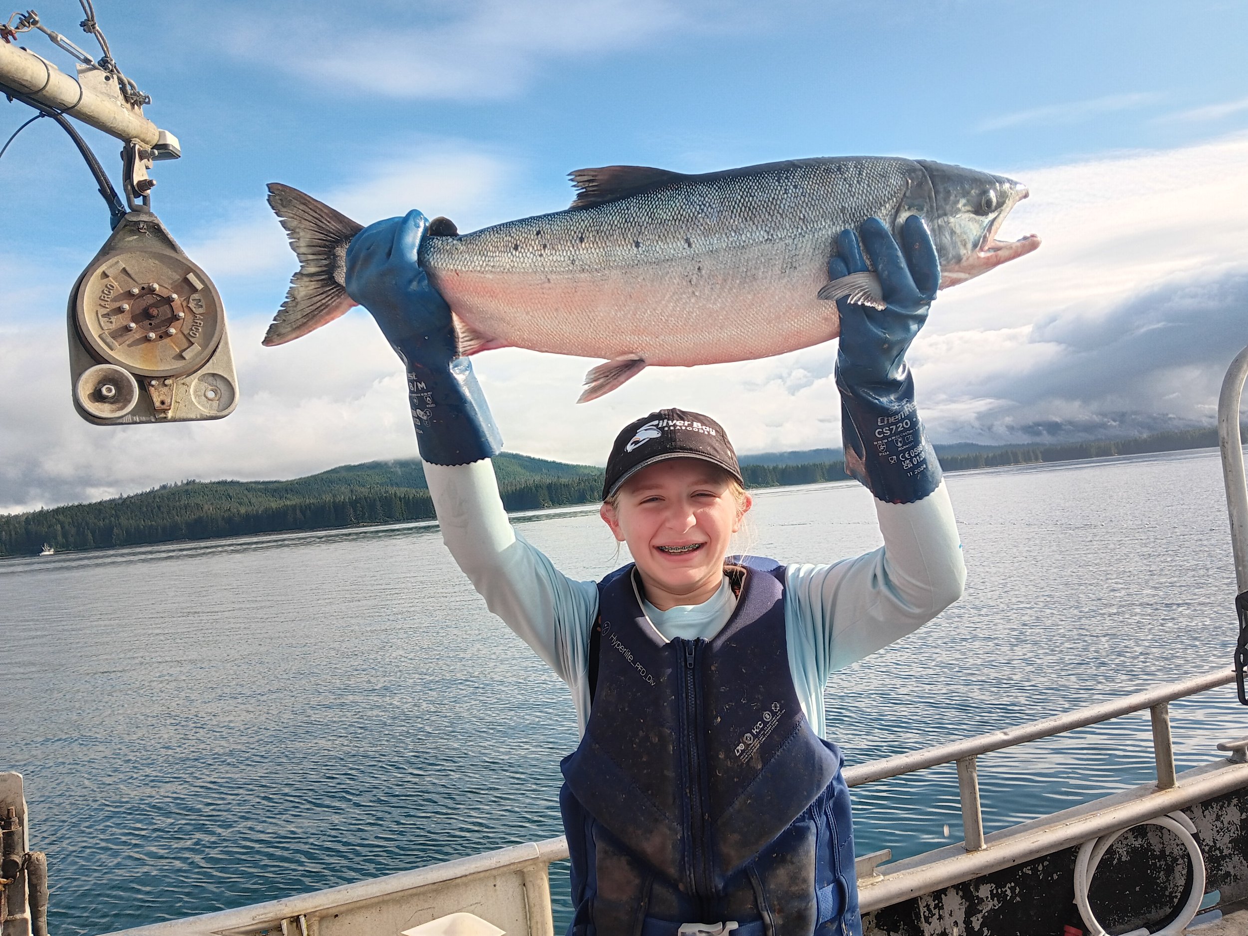 A young girl on a boat holding a large fish above her head with both hands, smiling, with a lake, mountainous landscape, and cloudy sky in the background.