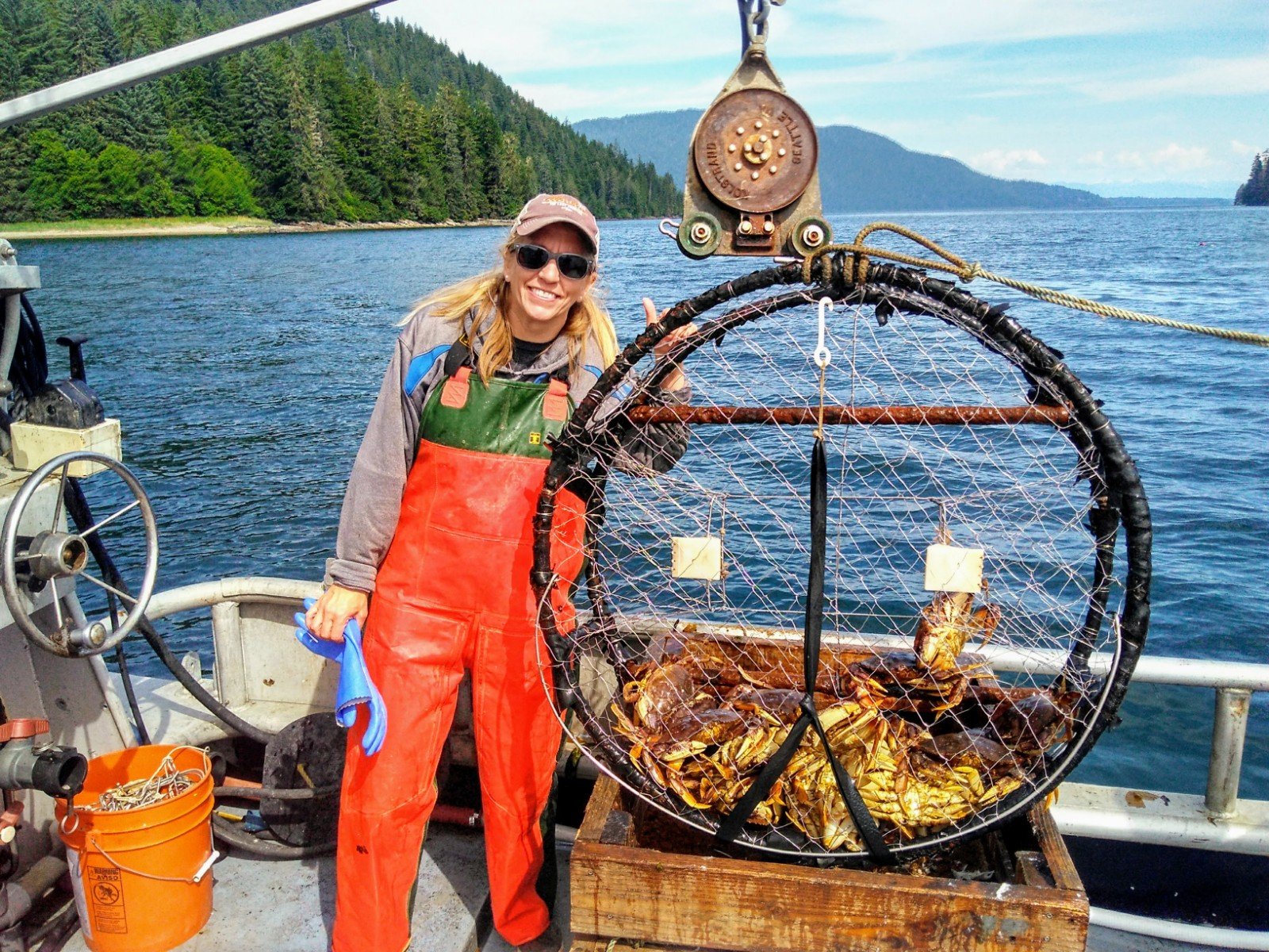 A woman in a red and green waterproof suit holding a crab, on a boat with a scenic water and forested mountain background.