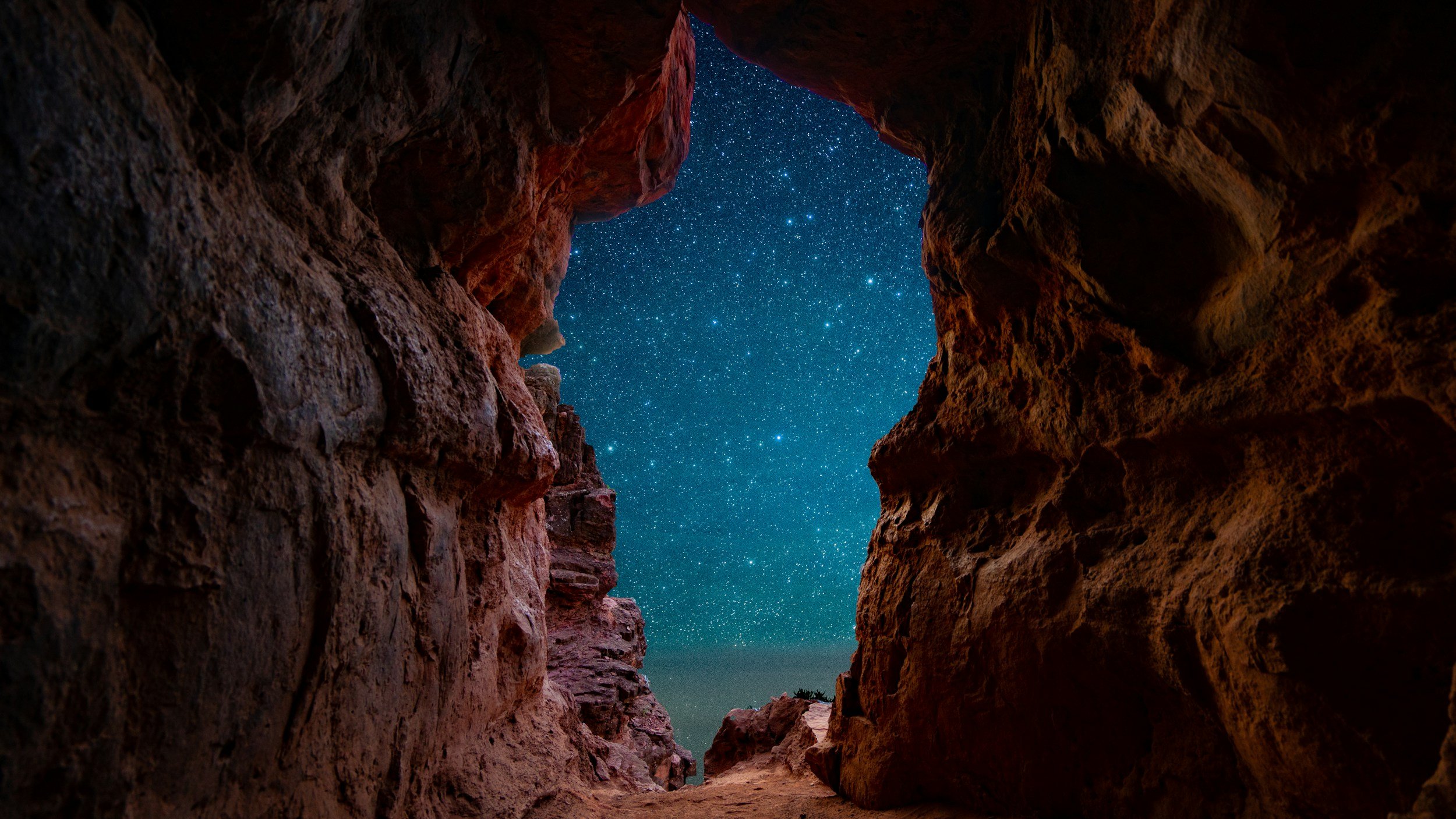 View of a starry night sky through the opening of a rocky cave or canyon with reddish-brown textured rock walls.