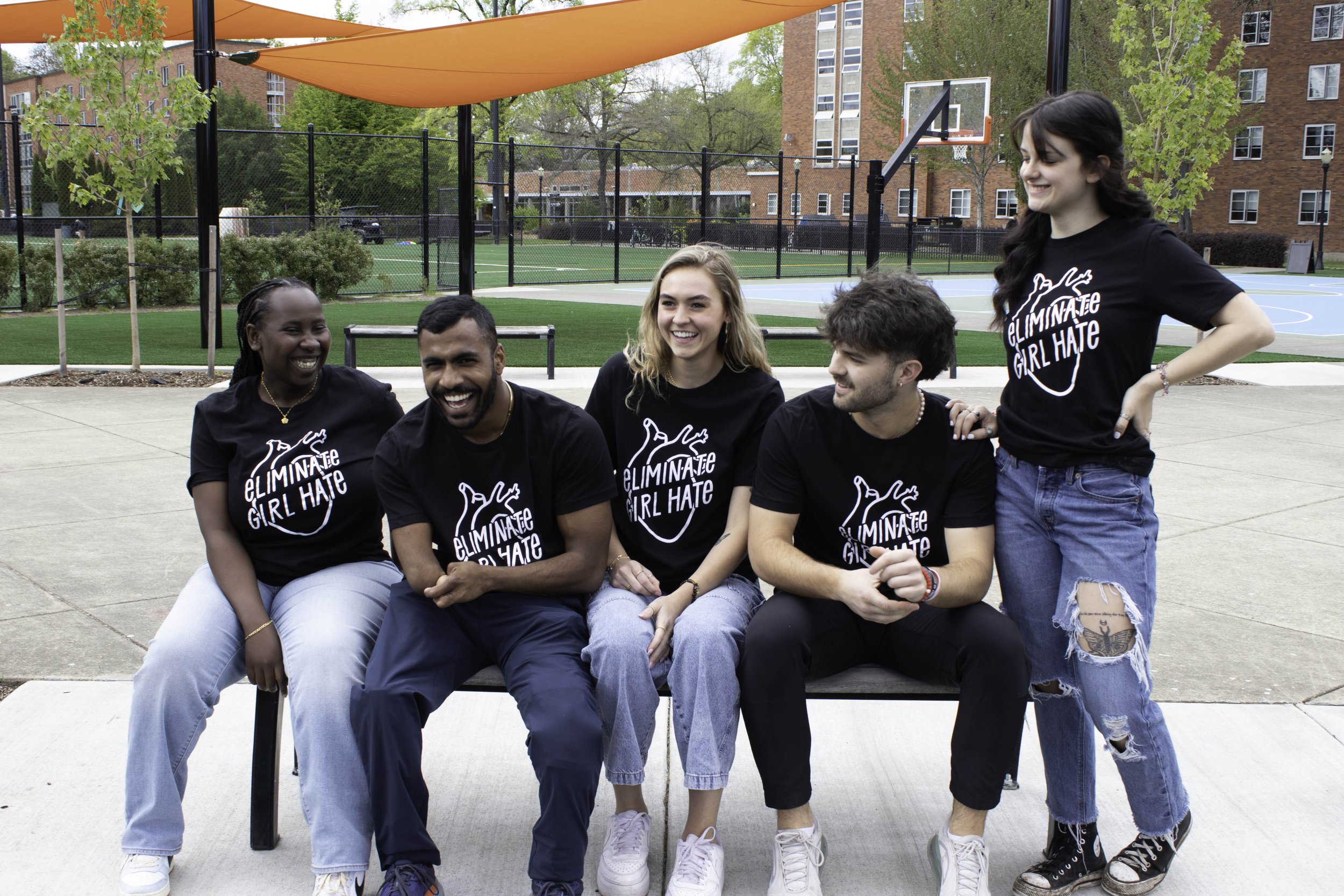 Group of college university students wearing jeans and black unisex Eliminate Girl Hate t-shirt while sitting on bench smiling and laughing