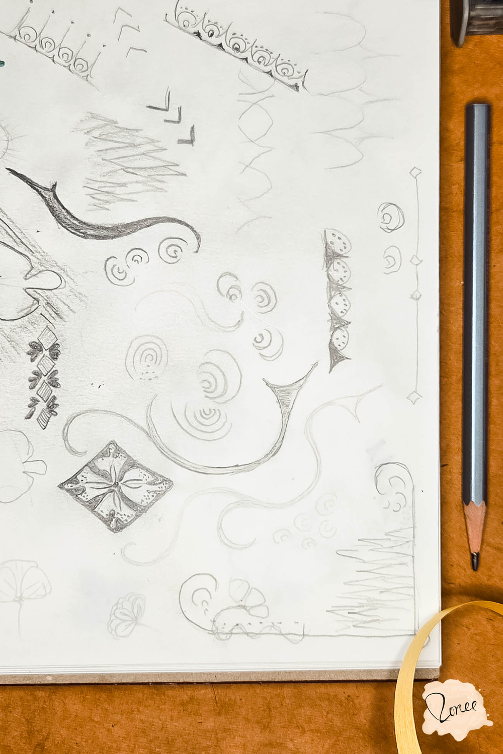 Close up flat lay image of a sketchbook with ornamental sketches,  a silver pencil is resting along the right,  and a yellow ribbon is curling into the frame from the bottom right..