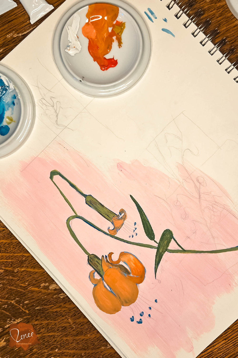 Flat lay image of a sketchbook with a gouache lily painting in progress and a ceramic palette dish with orange paint.
