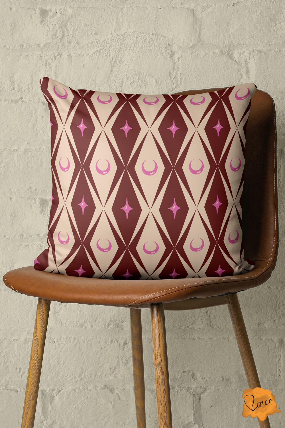 Throw pillow with burgundy and cream diamonds and pink moons and stars on a vintage wood and leather chair in front of a painted brick wall.