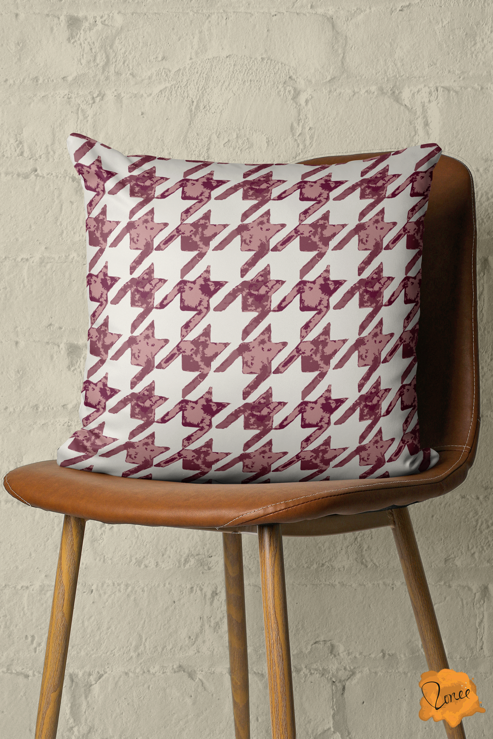 Throw pillow with rustic hand painted houndstooth in burgundy, mauve and off white sits on a vintage wood and leather chair in front of a painted brick wall.