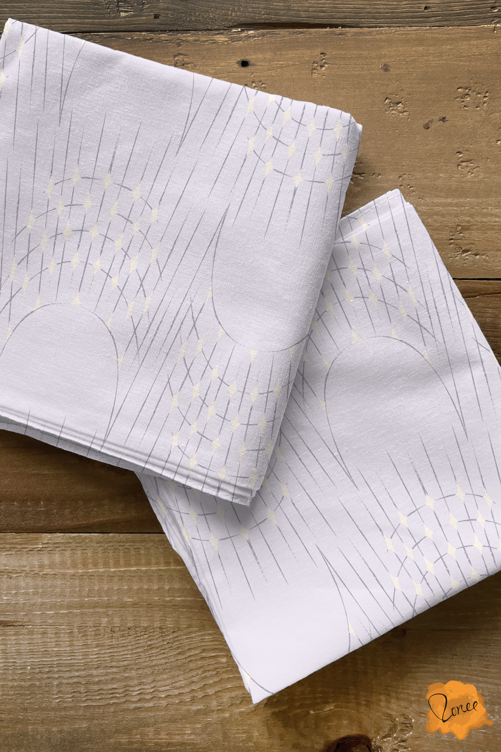 Two squares of folded quilting cotton with Delicate art deco arches and radisnt starburst lines with sparkling cream diamonds on pale lavender, rest on an old wood surface.