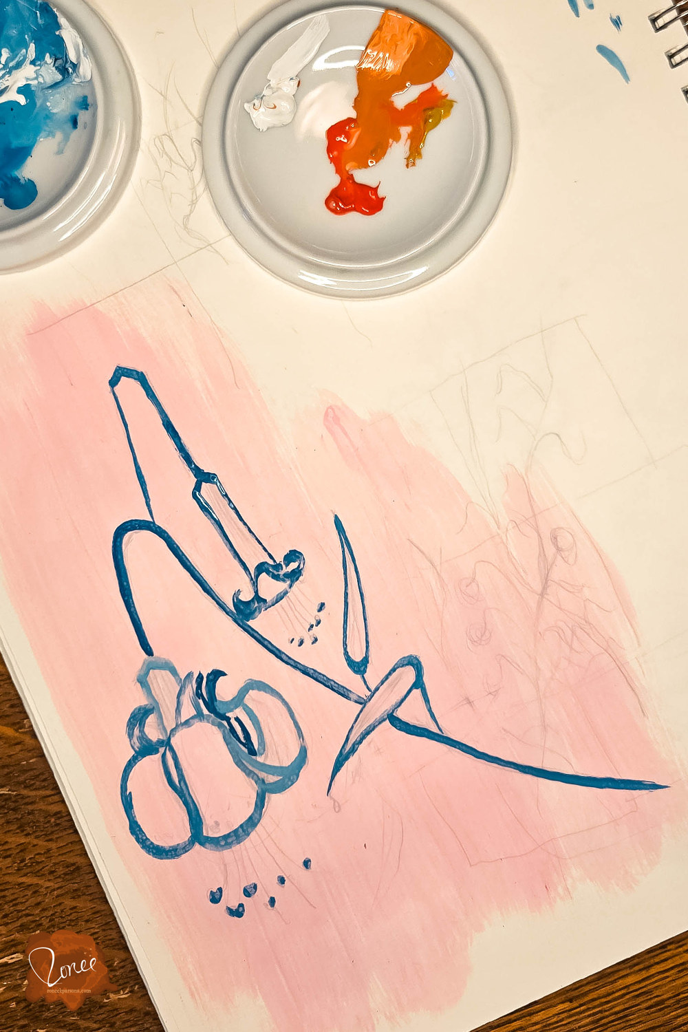 Sketchbook page with lilies and other marks sketched in pencil, a light pink wash over the top, and one lily outlined in blue paint that is still wet, two ceramic dishes sit on the pad one with orange and one with blue paint.