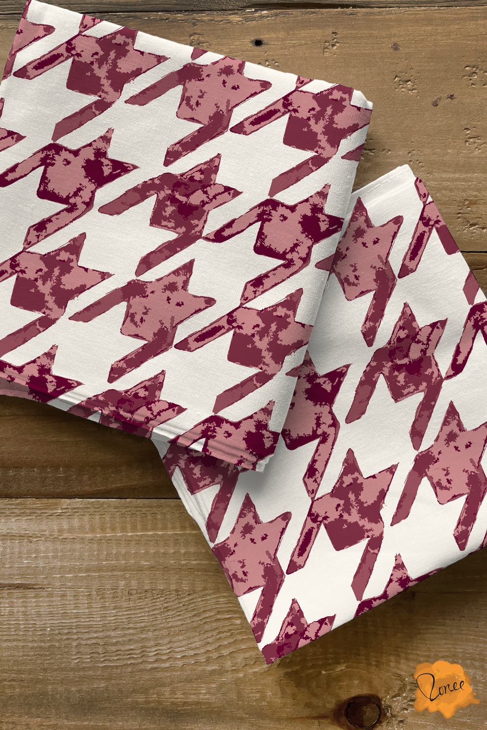 Two squares of folded quilting cotton with a rustic hand painted houndstooth pattern in deep burgundy and mauve on off white rest on an old wood surface.